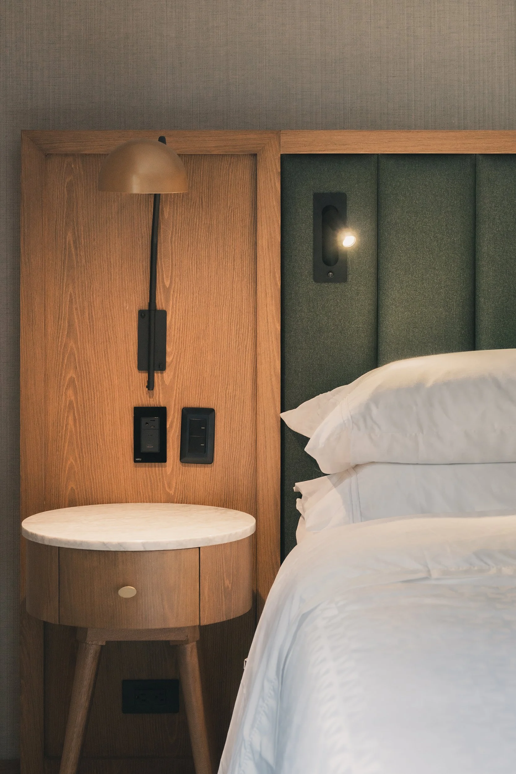 Close-up of a modern hotel room nightstand with a wooden surface, a small round marble table, and a bed with white linens and pillows. There is a wall-mounted light and outlet, with a dark green upholstered headboard in the background.