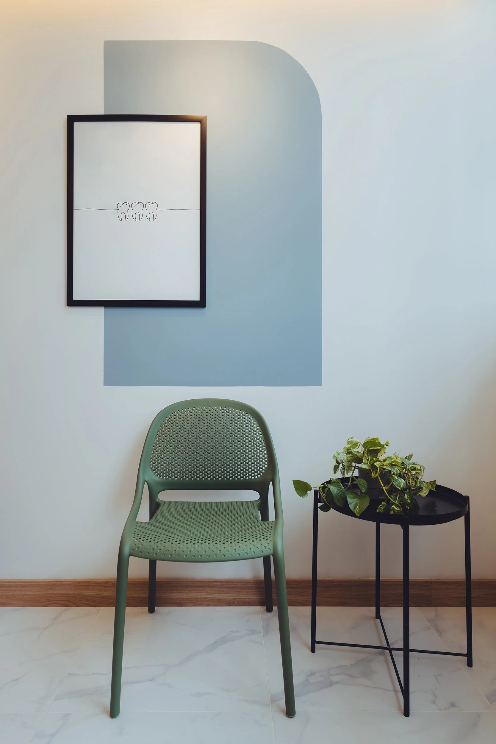 A mint green perforated plastic chair in front of a black round side table with a potted green leafy plant, positioned against a white wall with a framed minimalist line drawing of three teeth and a light blue area painted on the wall behind.