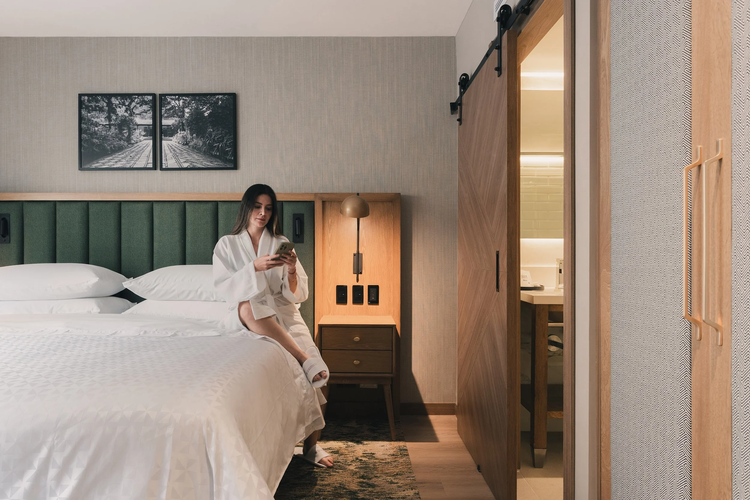 A woman in a white robe sitting on a bed, looking at her phone, in a modern hotel room with green upholstered headboard, framed black and white nature photographs, bedside lamp, and textured wallpaper.