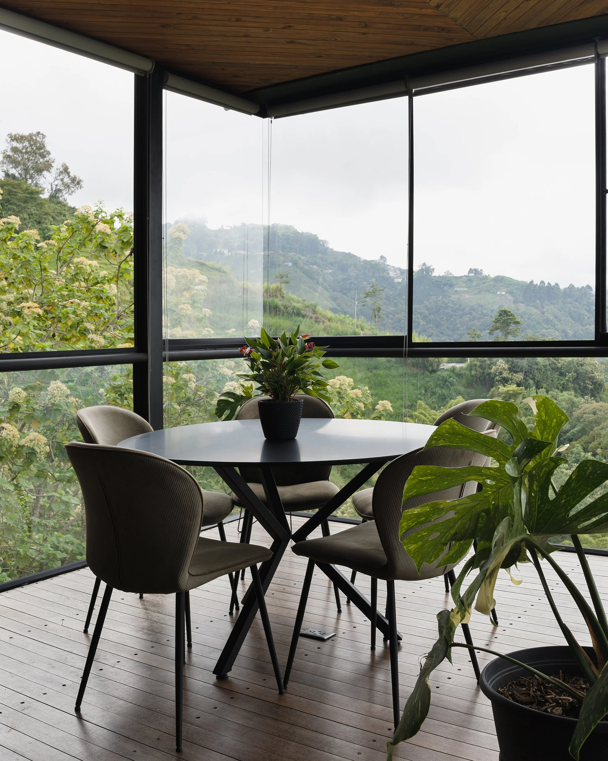 Fotografía de interiores de un comedor circular, con algunas plantas de interiores y en un espacio rodeado de ventanas piso techo. De fondo un paisaje de montañas y arboles