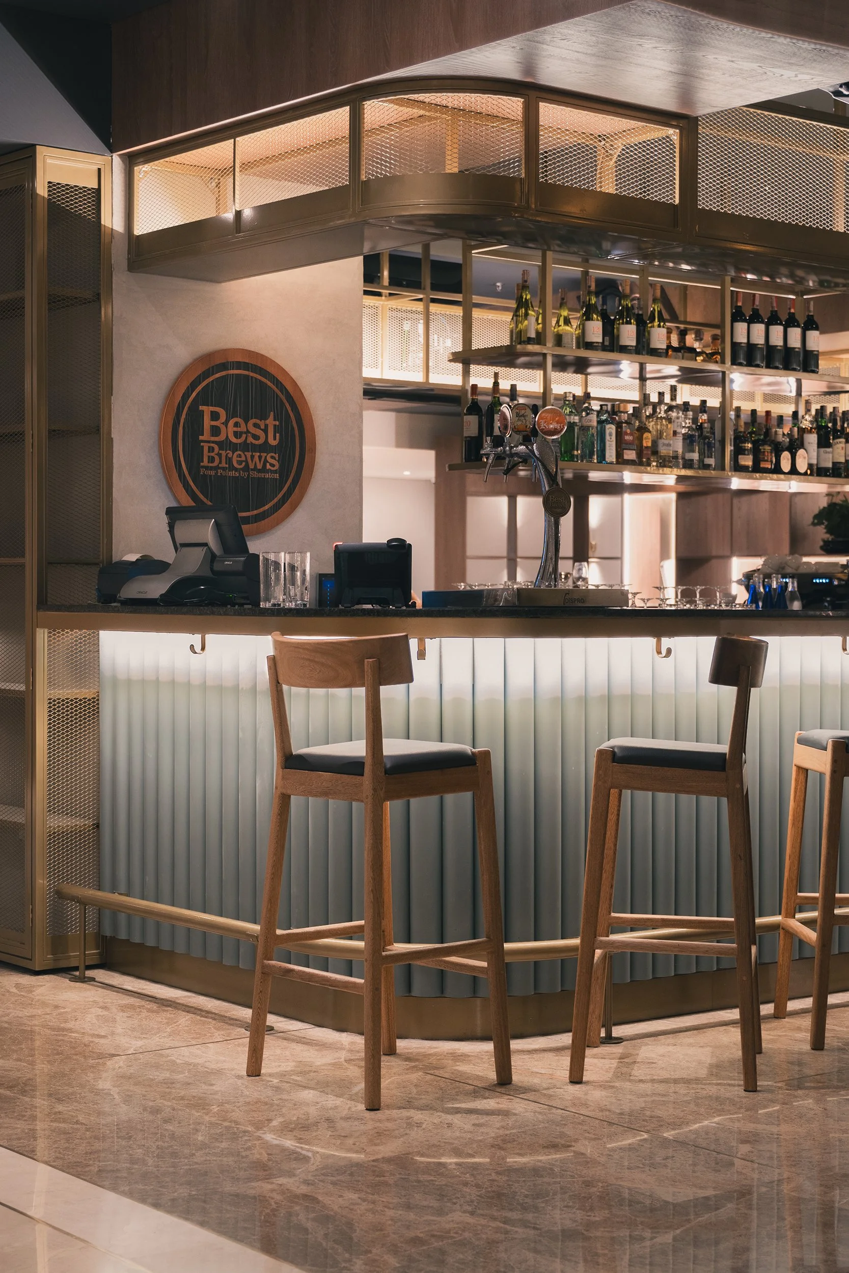 A bar area in a restaurant or hotel with wooden chairs and a counter with drinks. The sign on the wall says 'Best Brews, Four Points by Sheraton.' The bar has shelves with bottles of alcohol and a beer tap.