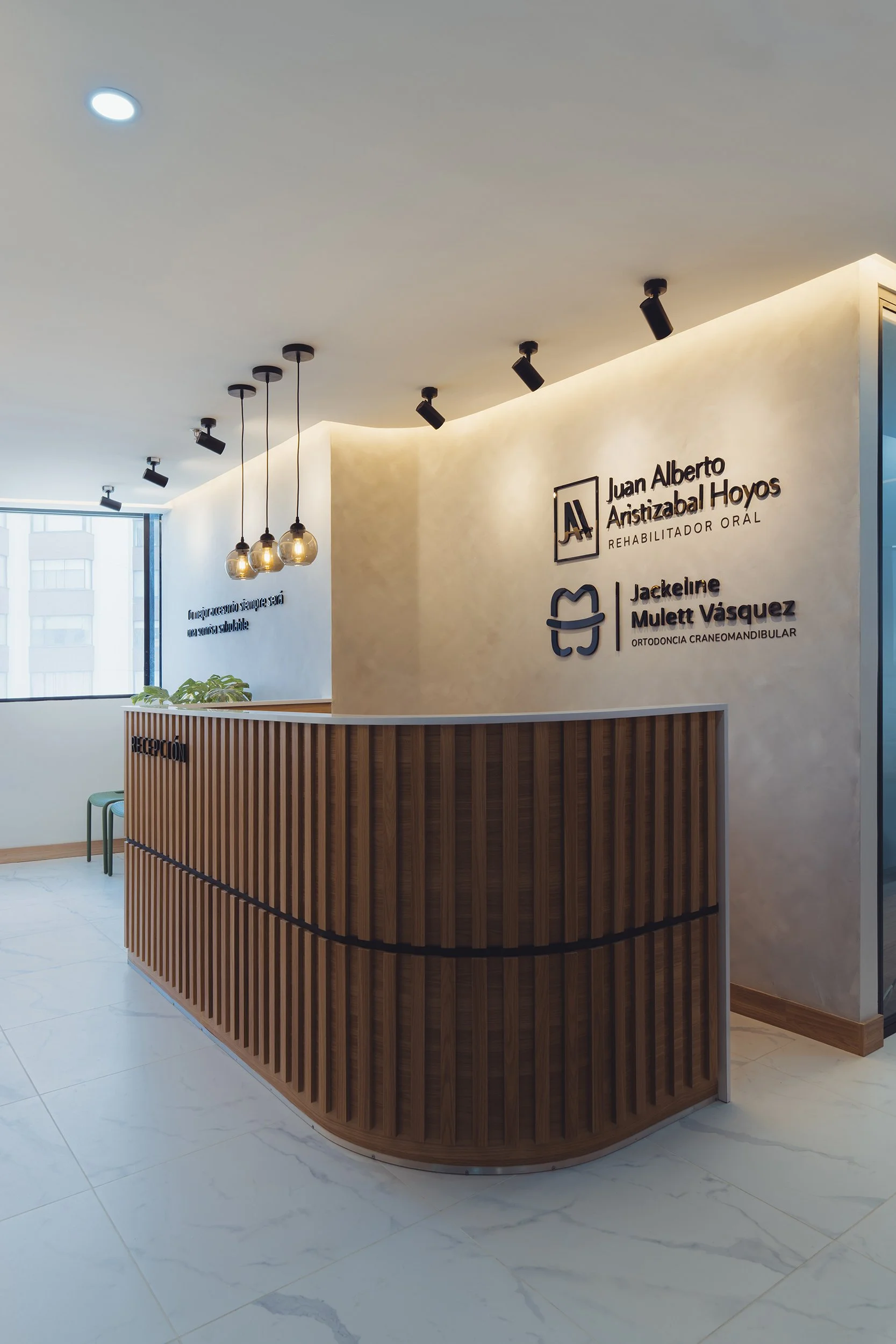 Reception area with a curved wooden desk, modern hanging light fixtures, a sign on the wall reading 'Juan Alberto Aristizabal Hoyos, Rehabilitador Oral,' and a window with city buildings outside.