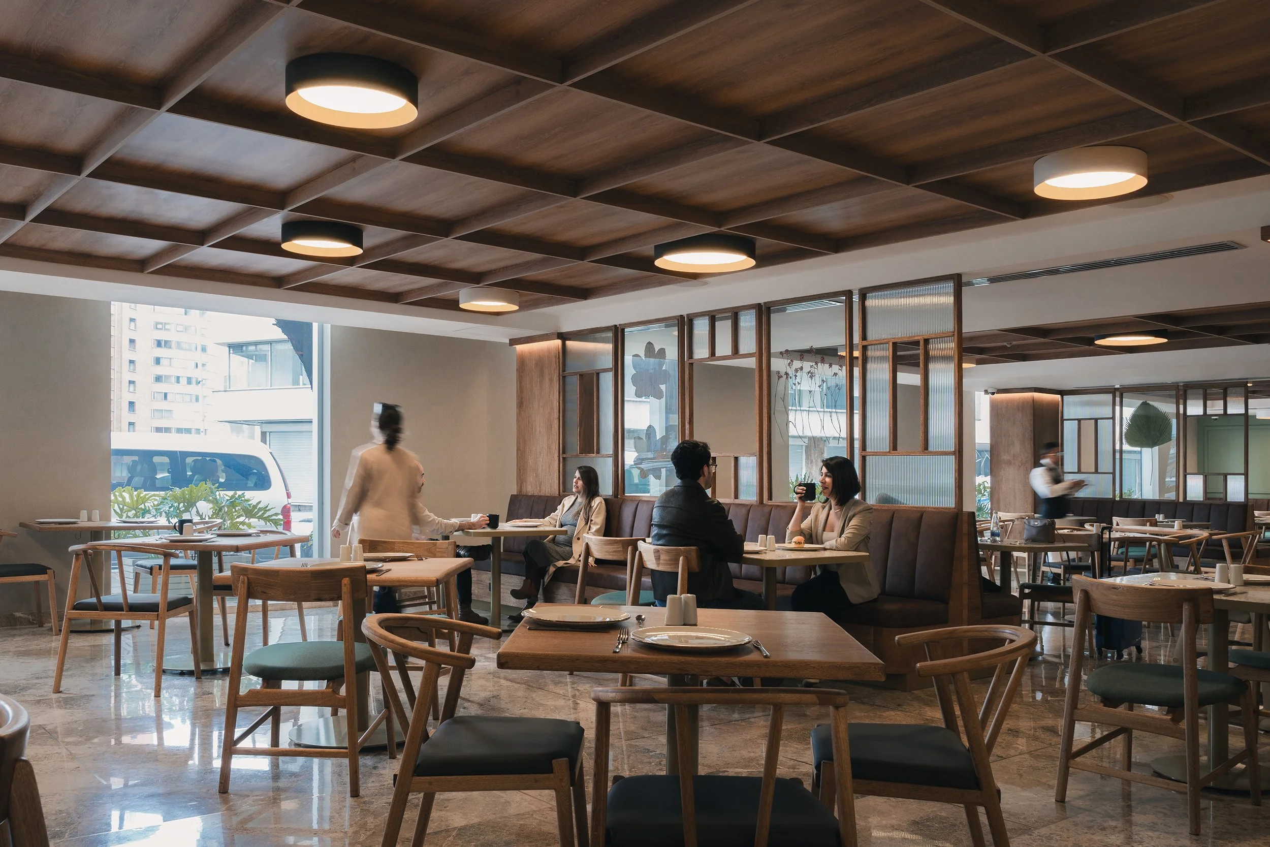 A modern restaurant interior with wooden tables and chairs, a wooden ceiling with recessed lighting, large windows showing city buildings outside, and a few customers dining and waiting staff serving.