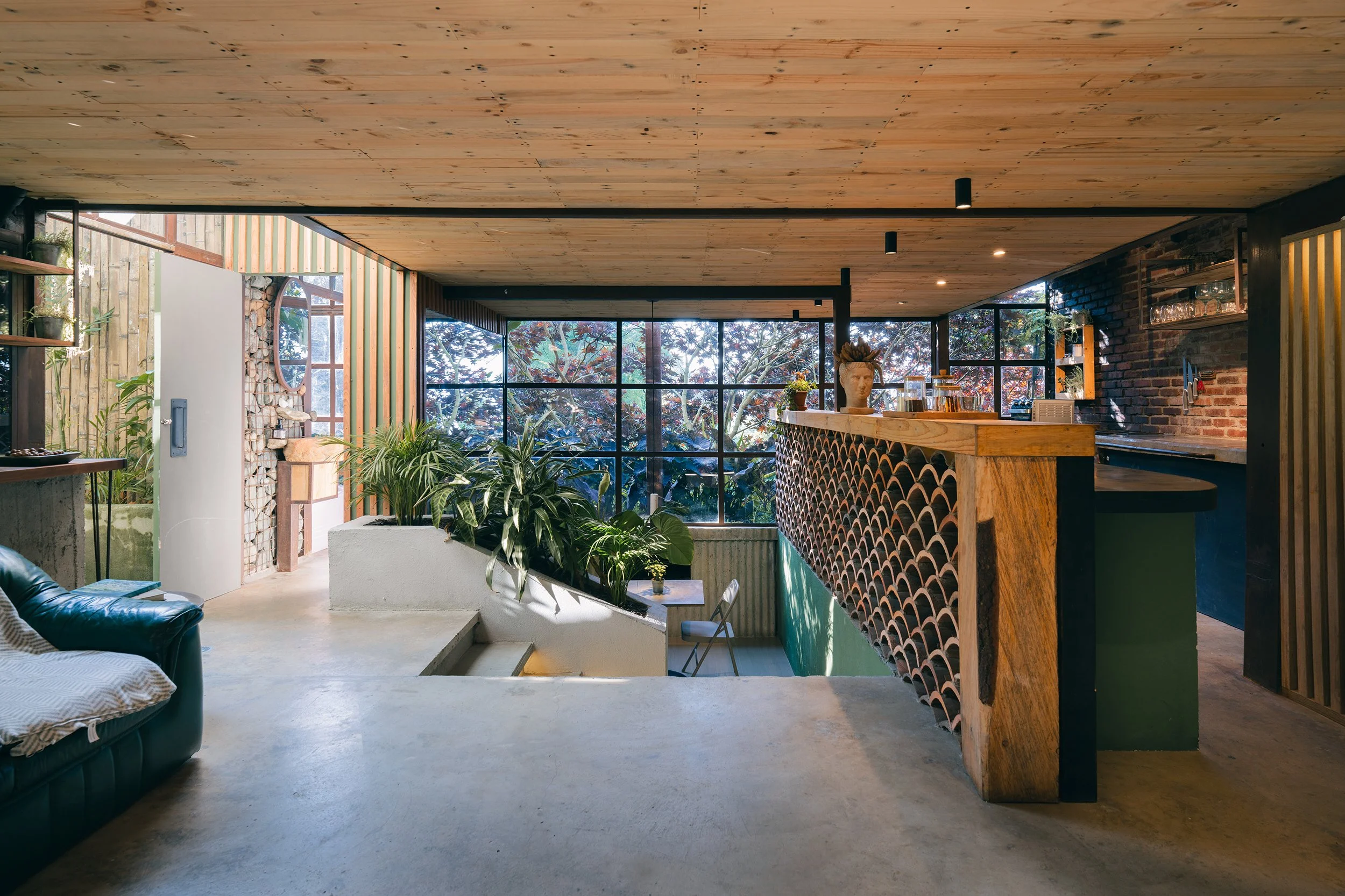 Interior of a modern house with a wooden ceiling, large windows showing trees outside, a bar area with decorative tiles, indoor plants, a sofa, and a white concrete staircase.