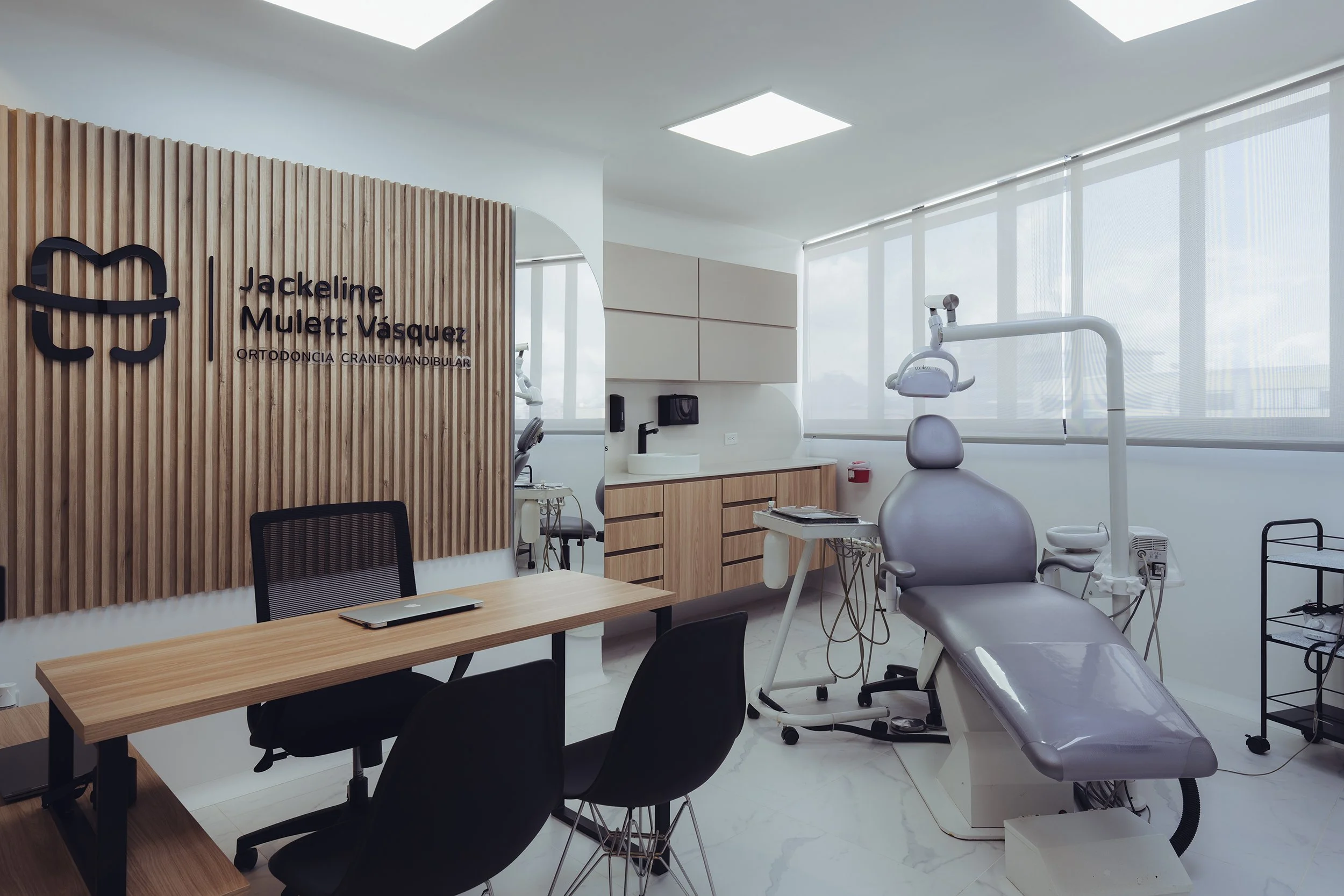 Interior of a modern dental clinic with a dental chair, dental instruments, a wooden reception desk, and wall signage reading 'Jackeline Mulet Vázquez, Orthodoncia Craneomandibular'. Large windows with blinds allow natural light to fill the space.