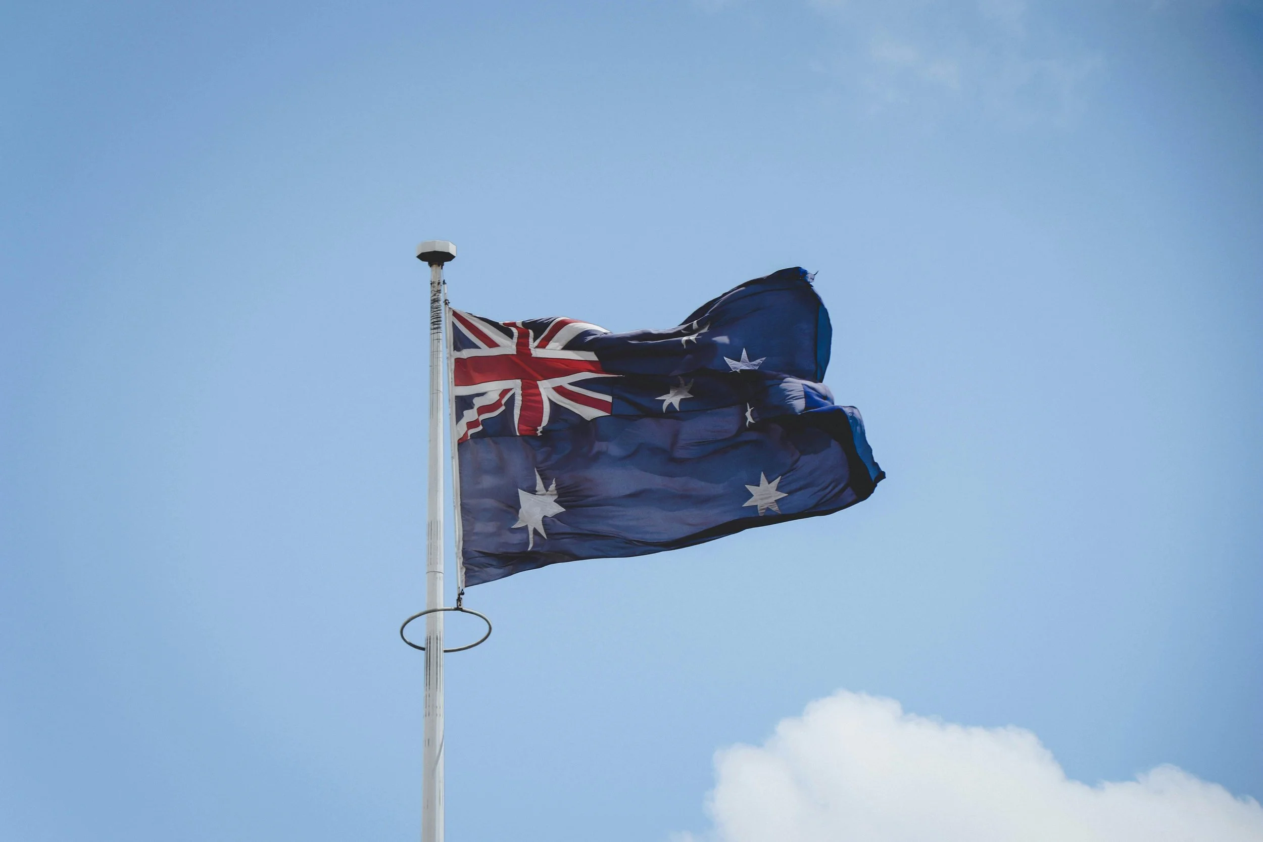 An Australian flag flying on a flagpole against a blue sky with some clouds.