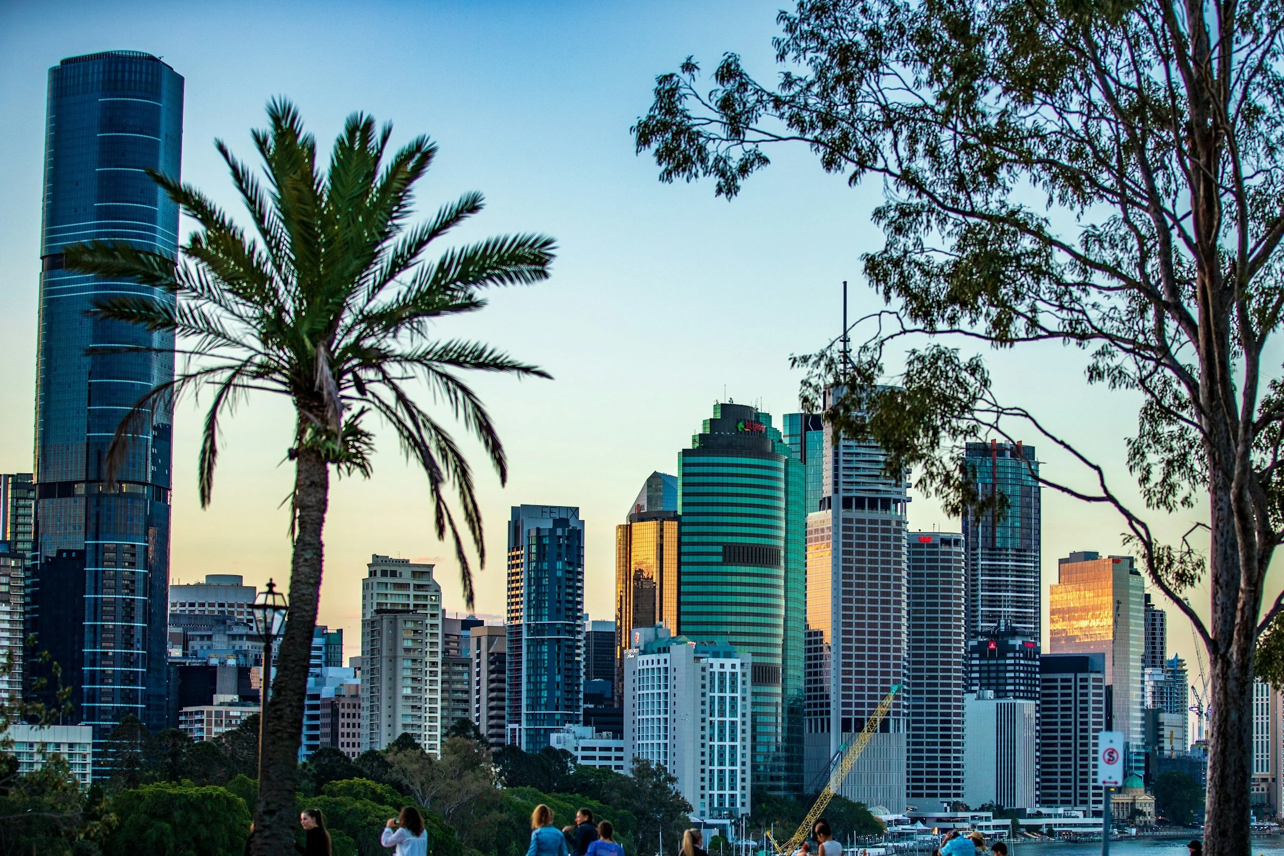 City skyline with tall skyscrapers, including a building with tinted glass, framed by palm trees and trees in the foreground, with people visible at the bottom.