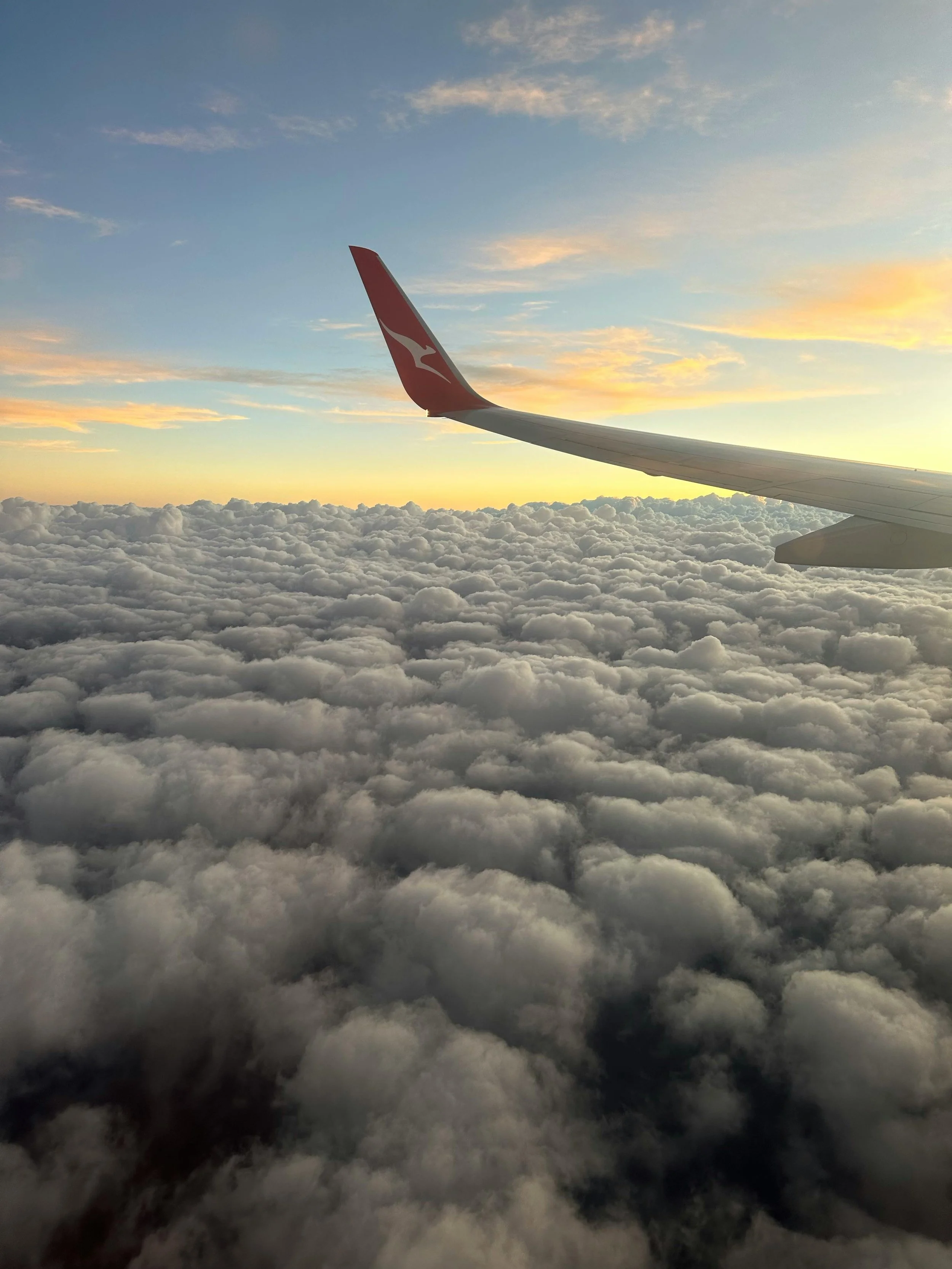 View from an airplane window showing clouds below and a wingtip with a red logo against a sky during sunset.