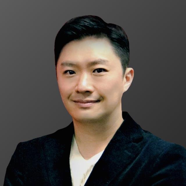 Portrait of an Asian man with short black hair, wearing a black blazer and white shirt, smiling slightly against a plain gray background.