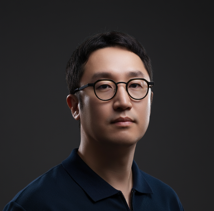 A young man with short black hair and glasses wearing a dark polo shirt against a dark background.