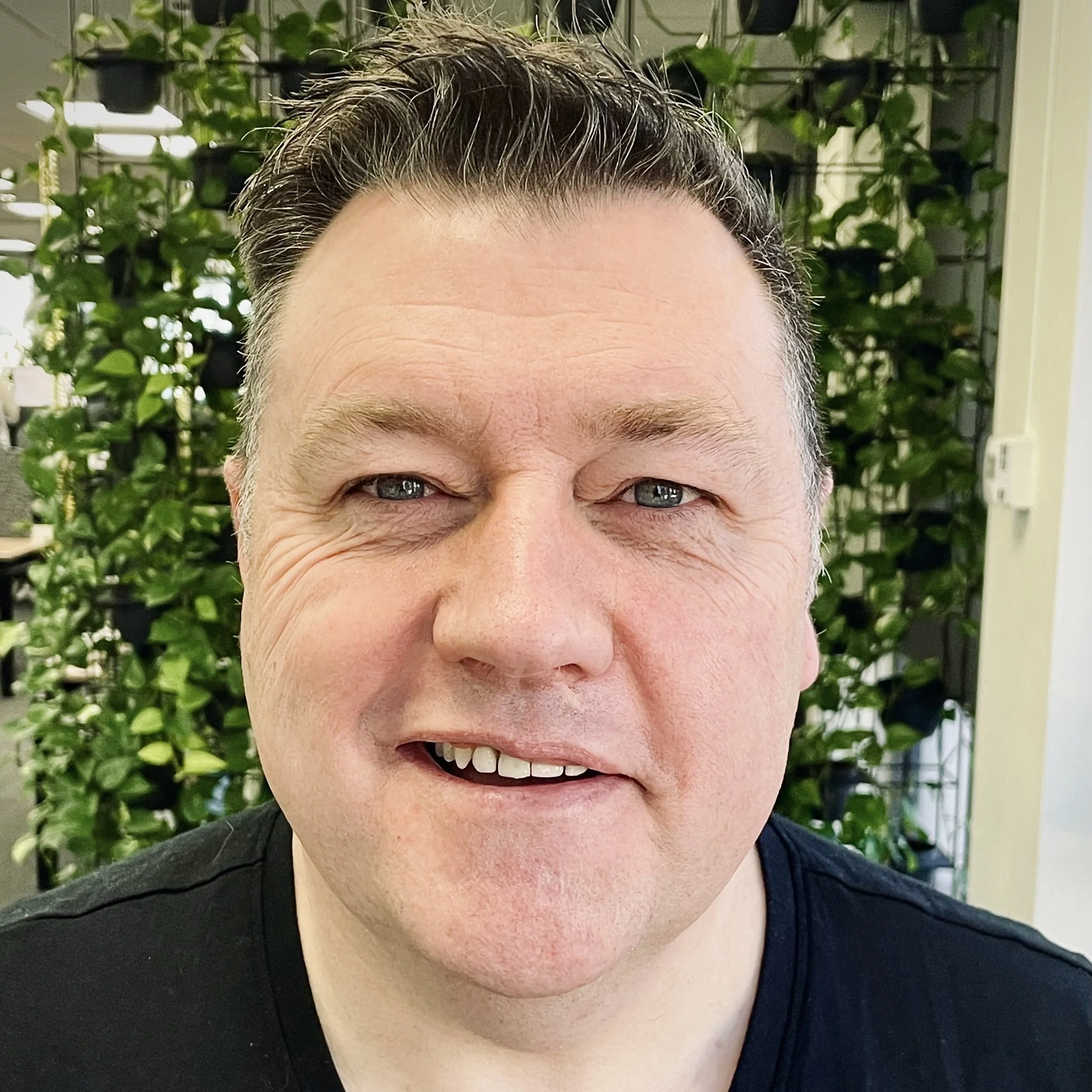 Close-up of a smiling man with short, dark, slicked-back hair, wearing a black shirt, with a background of green leaves and plants.