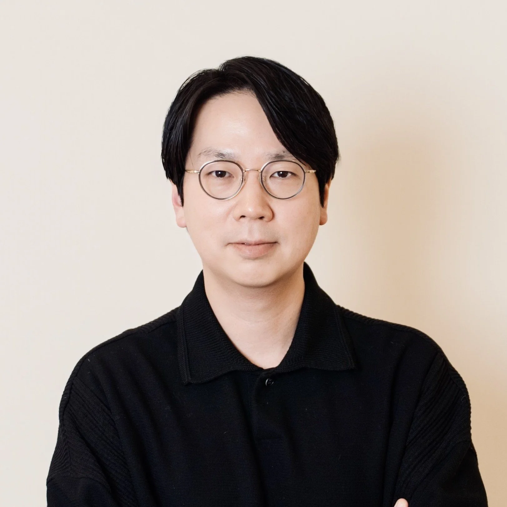 A young man with black hair, glasses, and a black shirt, smiling and posing against a light gray background.