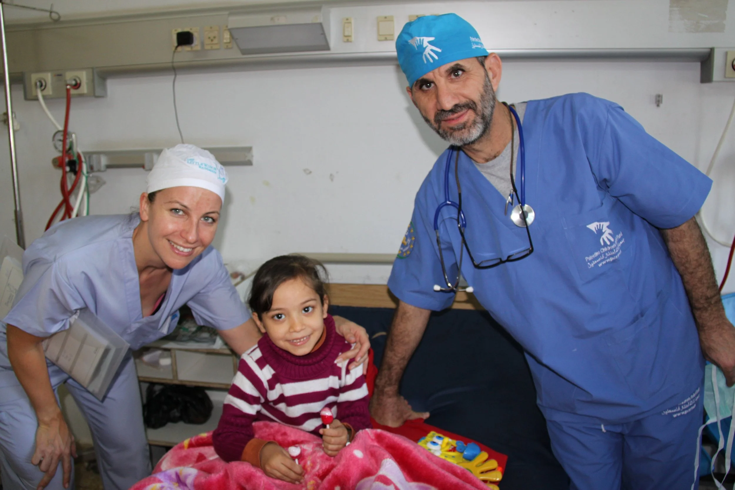 A doctor and a nurse with a young girl in a hospital room, smiling. The girl is sitting on a bed with a colorful blanket, holding a lollipop. The healthcare professionals are wearing scrubs and medical caps.