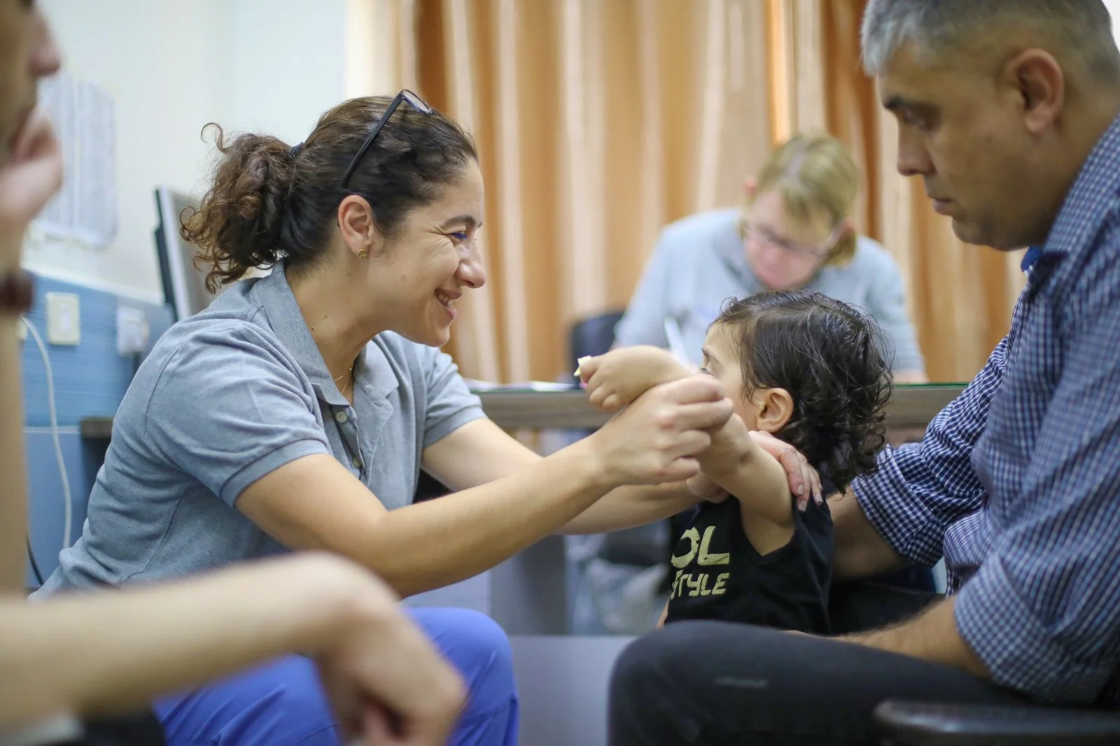 A woman smiling and playing with a young child while a man holds the child in a medical setting.