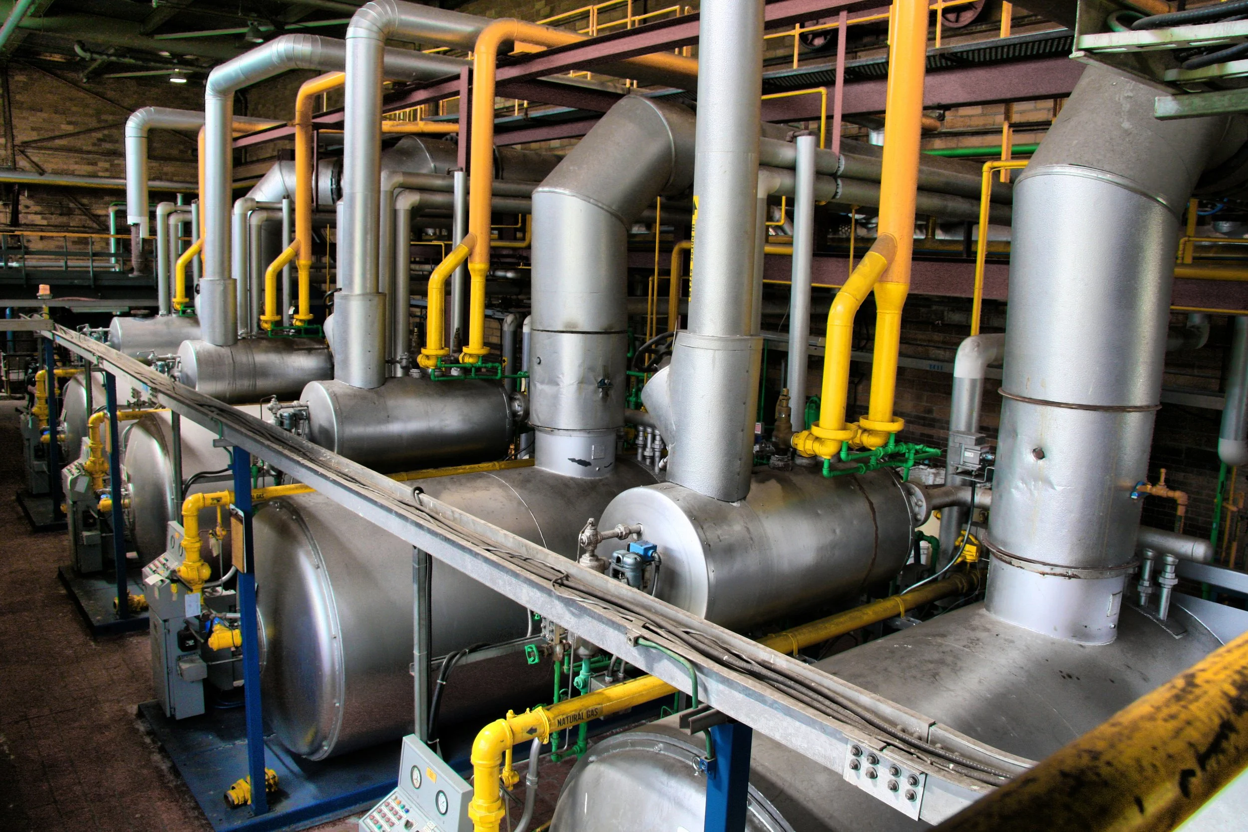 Industrial machinery with large metal tanks, pipes, and valves, including yellow and green components, inside a factory setting.