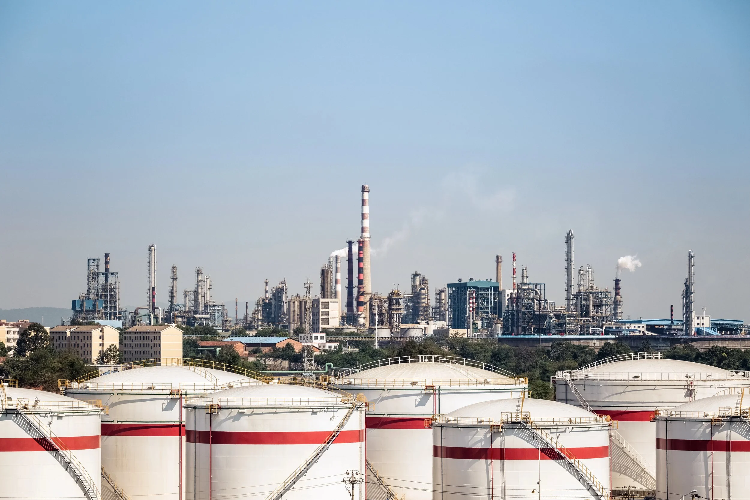 Industrial oil refinery with large storage tanks in the foreground and refinery processing units and smokestacks in the background.
