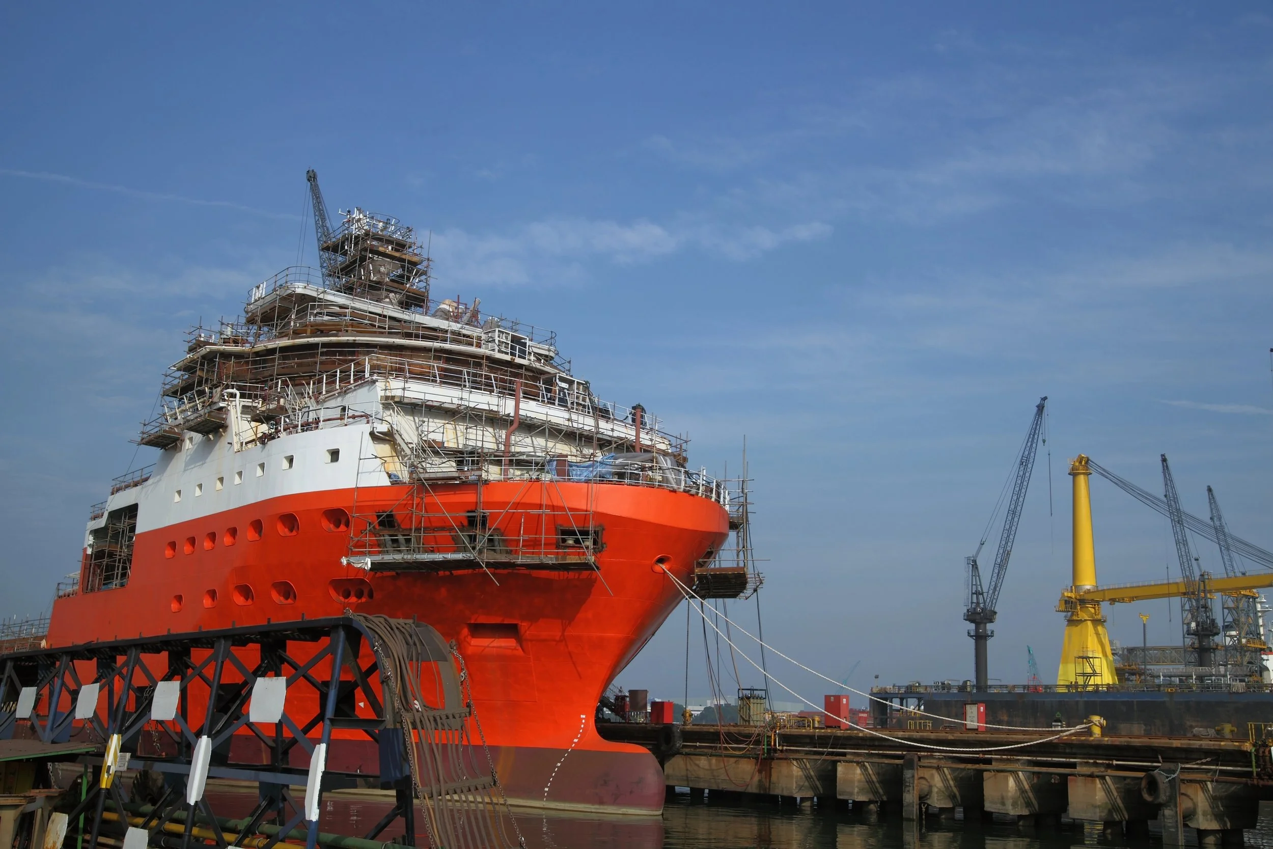 A large ship being constructed or maintained at a shipyard, with scaffolding around it and cranes in the background, under a partly cloudy sky.