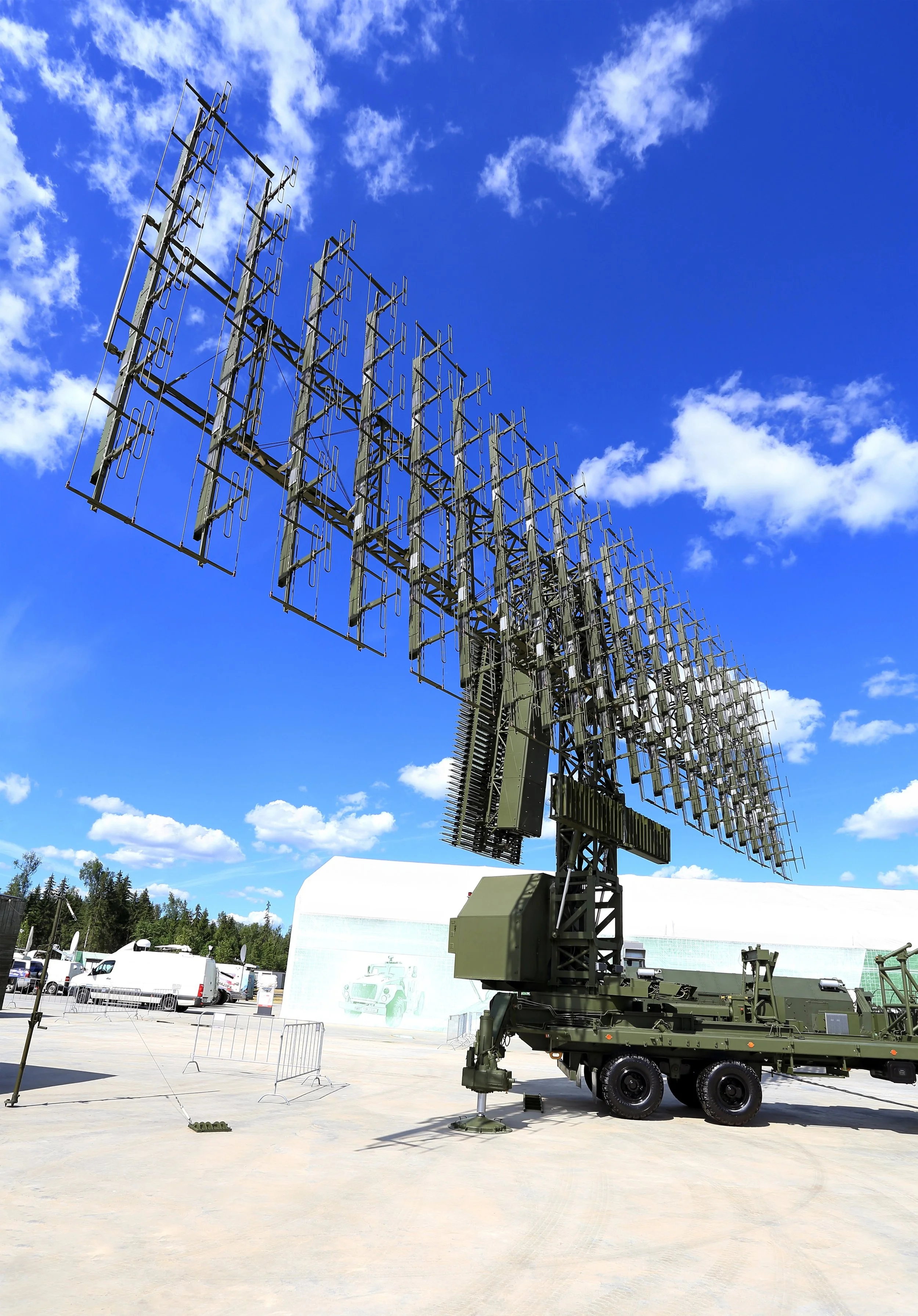 A large military radar antenna with multiple panels mounted on a wheeled platform outdoors, under a bright blue sky with scattered clouds.