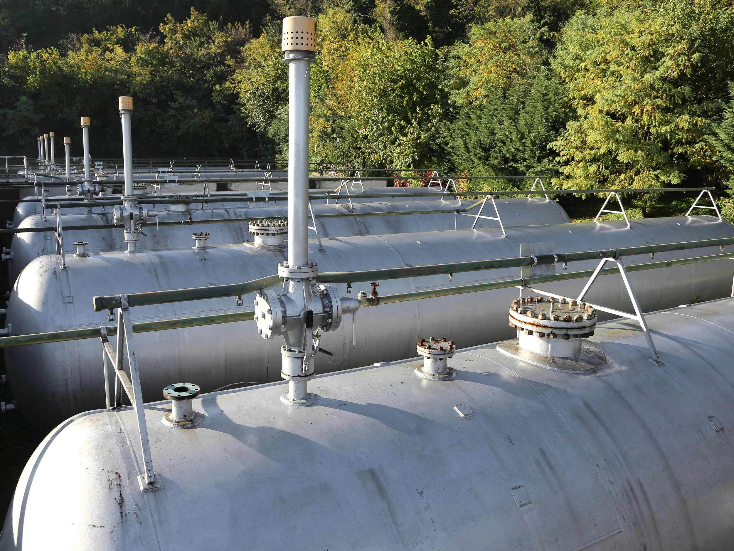 Multiple large metal storage tanks outdoors with green trees in the background.