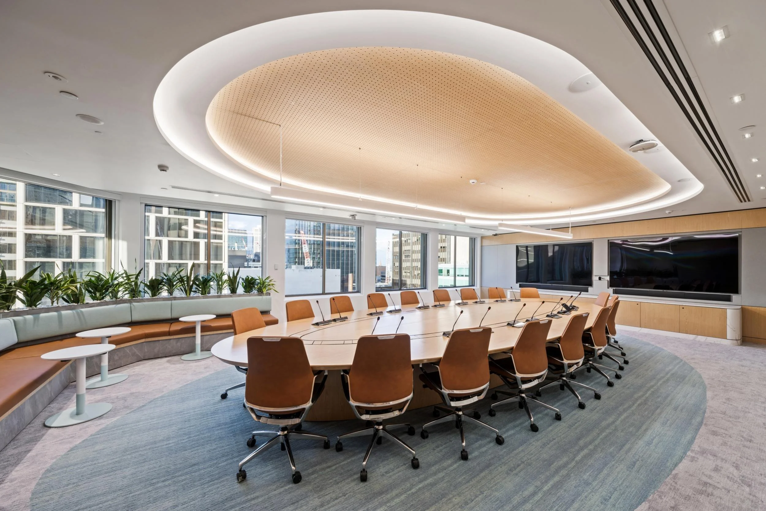 Modern conference room with large oval table, brown chairs, large windows, and a wall-mounted screen, decorated with indoor plants.