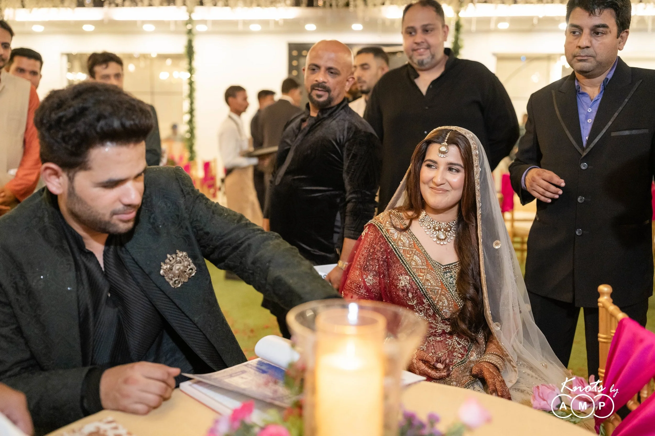 A group of people at a wedding reception, with a woman in traditional Indian attire sitting at a table, surrounded by men in formal wear.