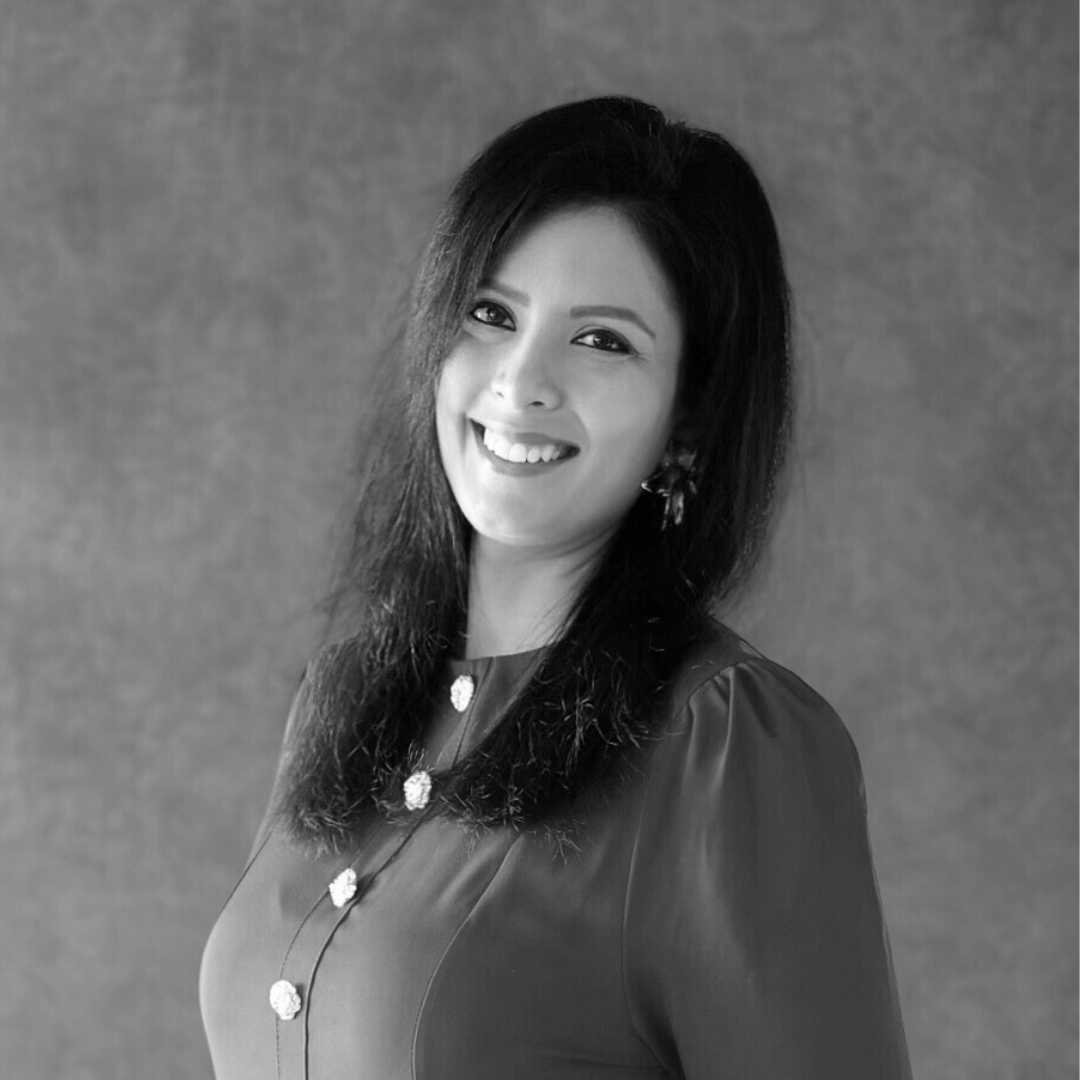 Black and white portrait of a woman smiling, with long dark hair, wearing earrings and a necklace, against a plain background.