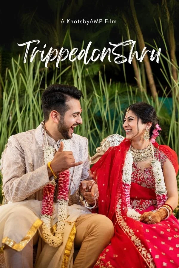 A groom and bride in traditional Indian wedding attire sitting outdoors on a decorated stage, smiling and enjoying each other's company, surrounded by green plants. The groom is giving a thumbs-up gesture.