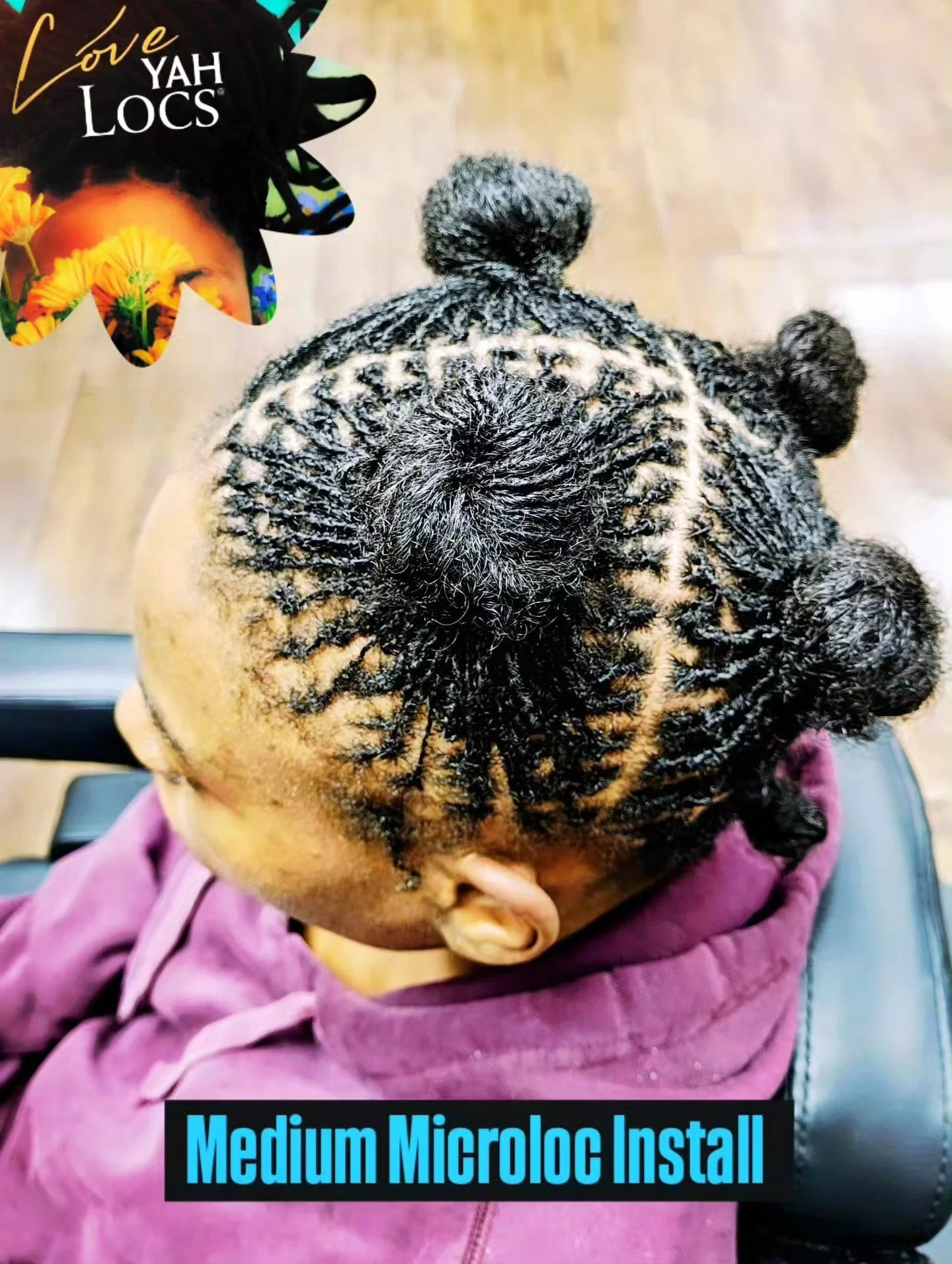A person with intricately styled medium-sized micro locs in a patterned updo, seated in a chair, wearing a purple hoodie, viewed from above.
