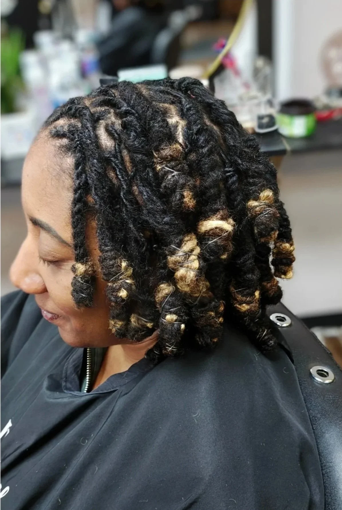 Close-up of a woman with freshly styled dreadlocks that are black with blonde tips, sitting in a salon chair with a black cape, hair tools and products in the background.