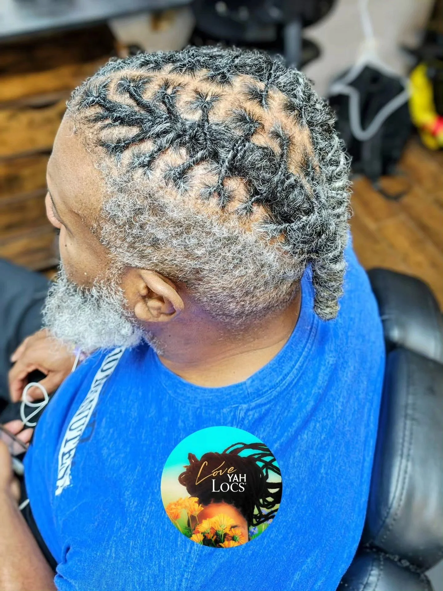 Top view of a man's head with gray and black dreadlocks, a gray beard, and wearing a blue shirt with a circular sticker that says 'Love Yah Locs' and features a woman with dreadlocks.