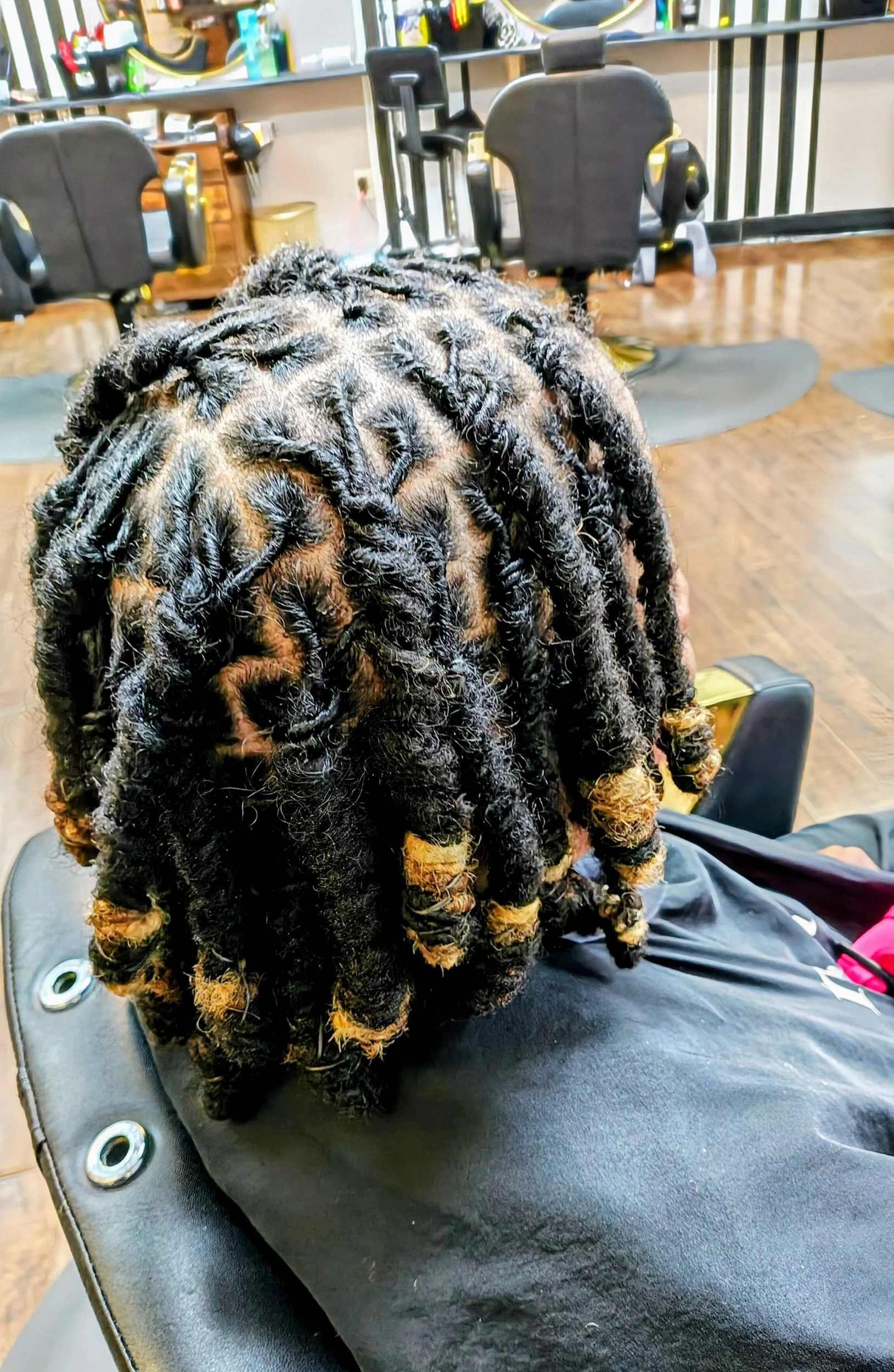 Close-up of a woman with freshly styled dreadlocks that are black with blonde tips, sitting in a salon chair with a black cape, hair tools and products in the background.