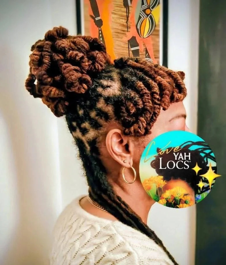 Side profile of a woman with styled brown and black locs, some in a bun on top, wearing hoop earrings and a cream sweater, standing indoors near a colorful abstract painting.