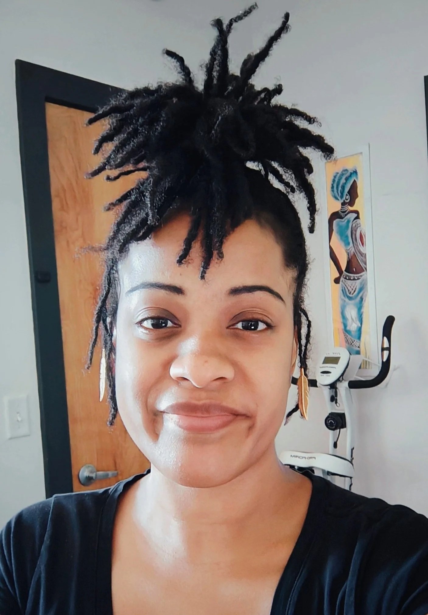 A woman with styled dreadlocks taking a selfie in a room with a painting of an African woman in traditional attire and an exercise machine in the background.