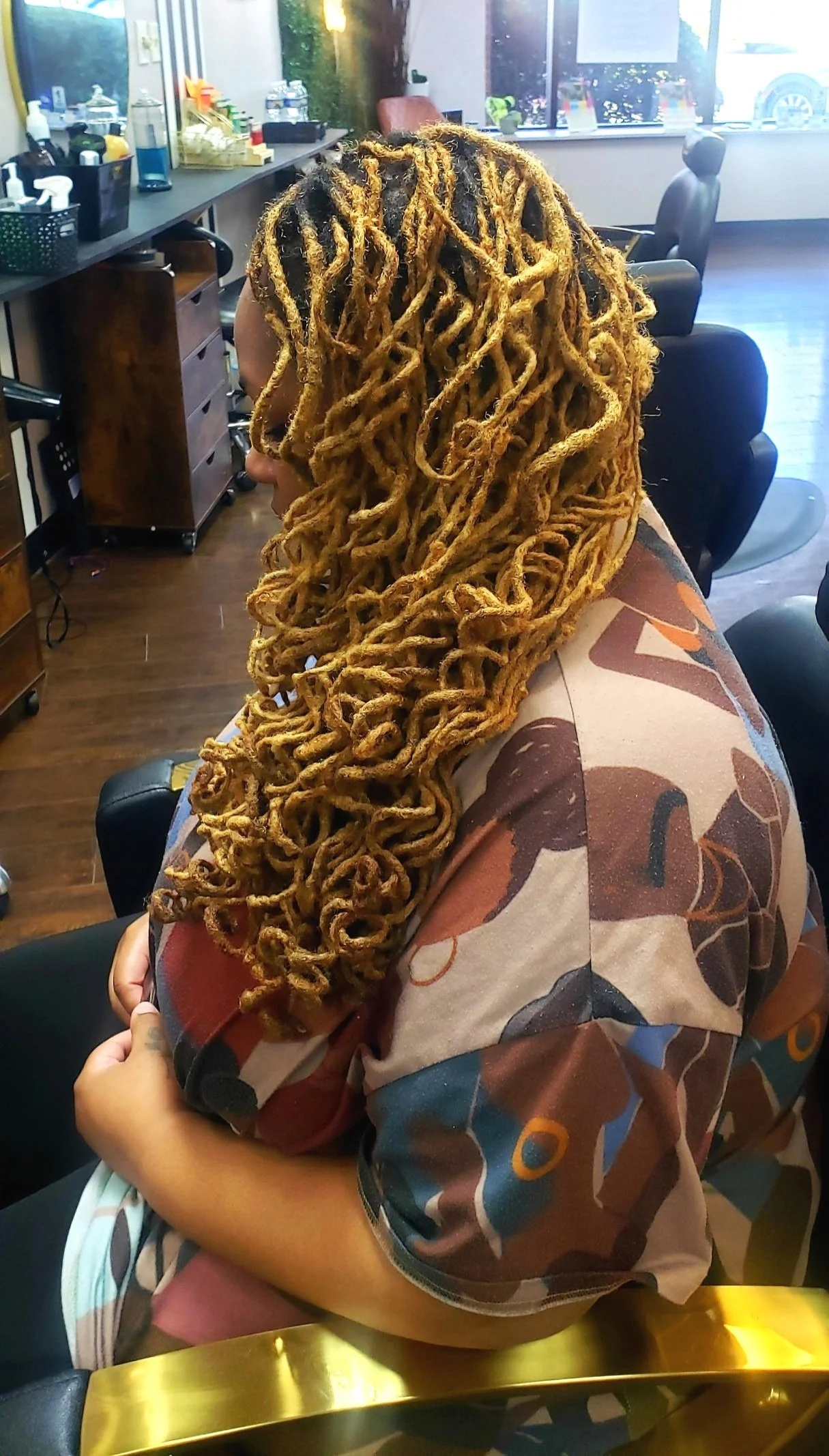 A person with long, blonde dreadlocks sitting in a salon chair, wearing a patterned shirt.