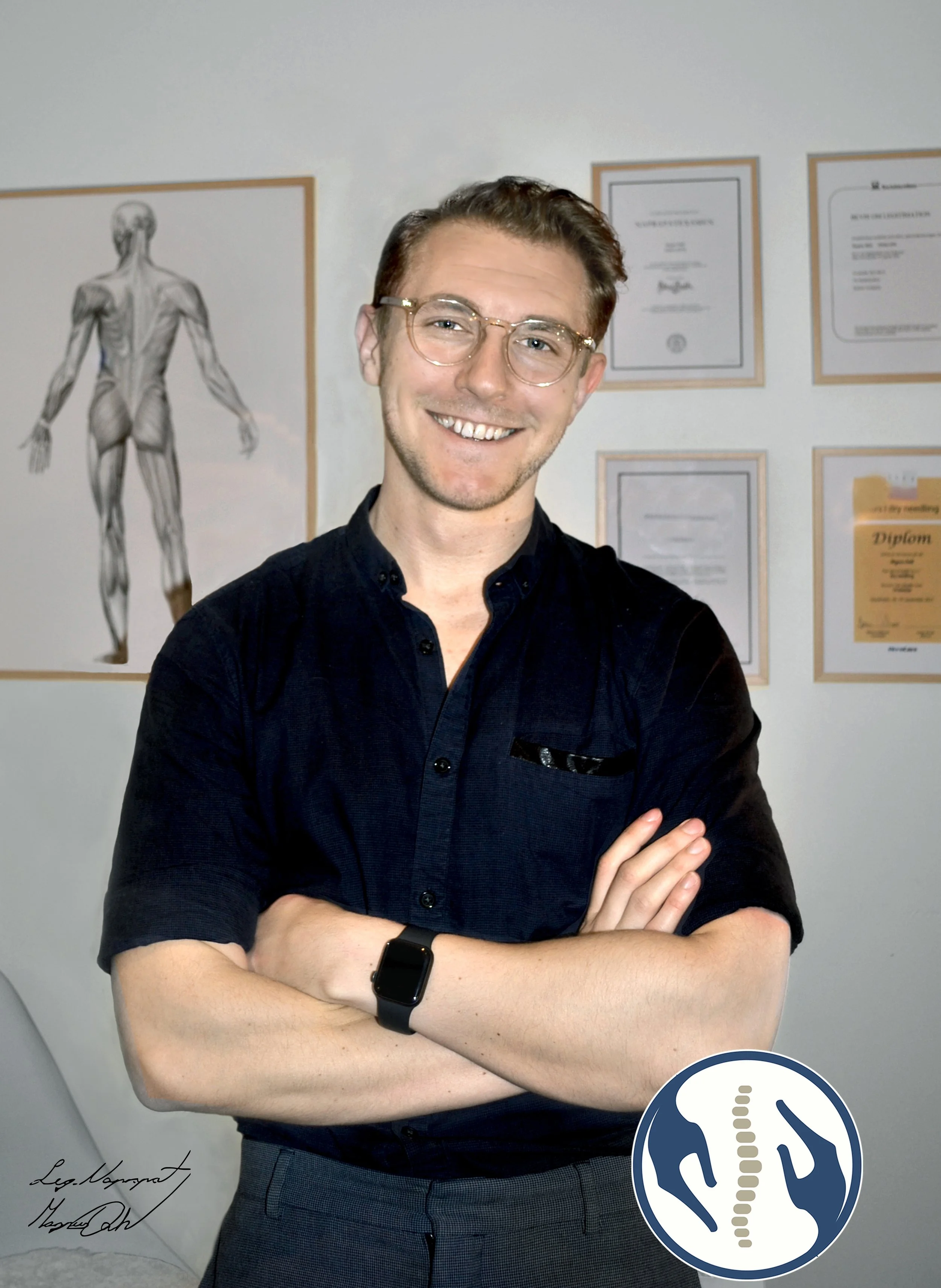 Person i mörk skjorta med glasögon som ler framför anatomiska affischer och diplom på en vägg.