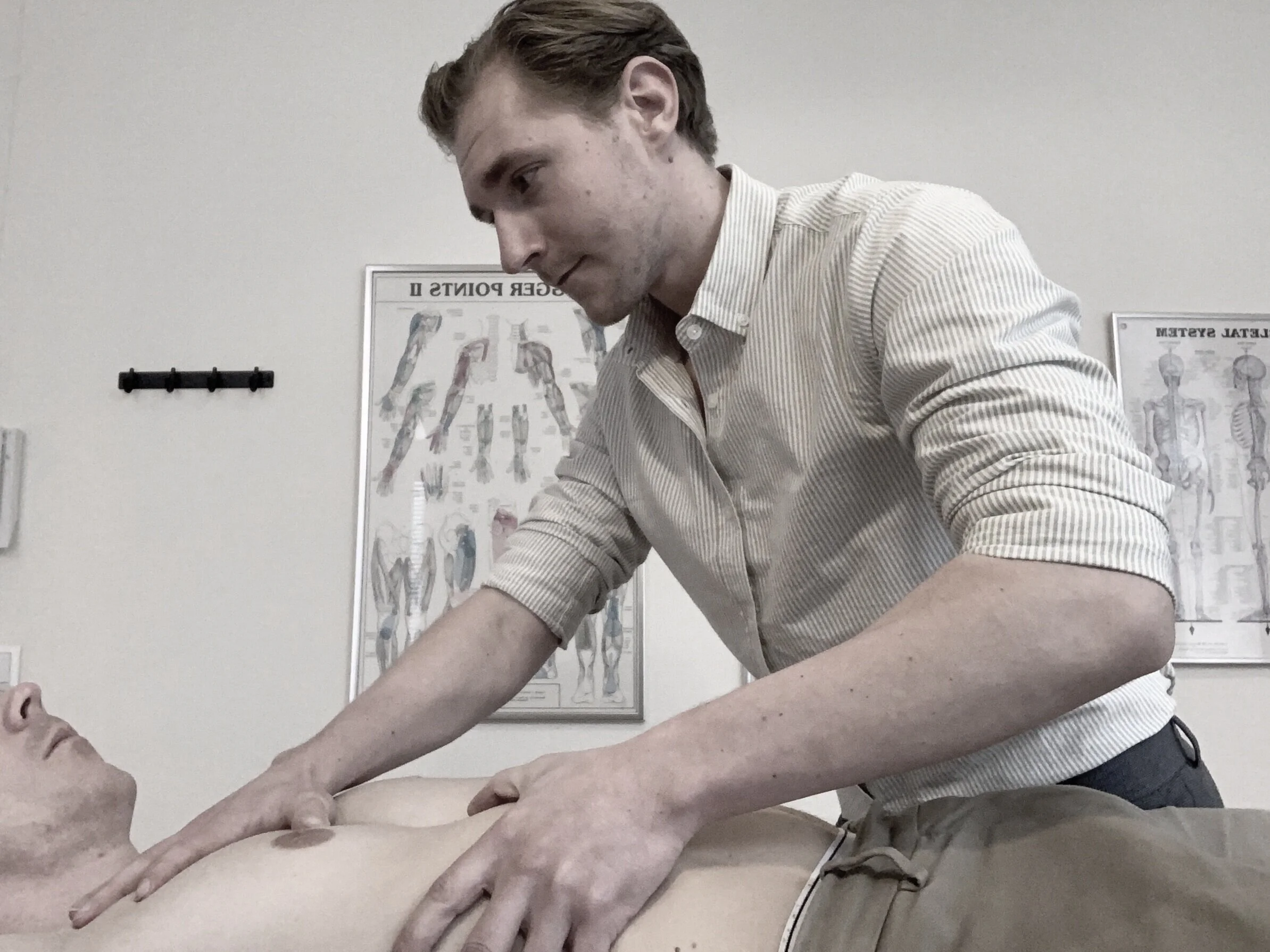 En person utför en medicinsk undersökning eller behandling på en annan person i en klinikmiljö, med anatomiska affischer i bakgrunden.
