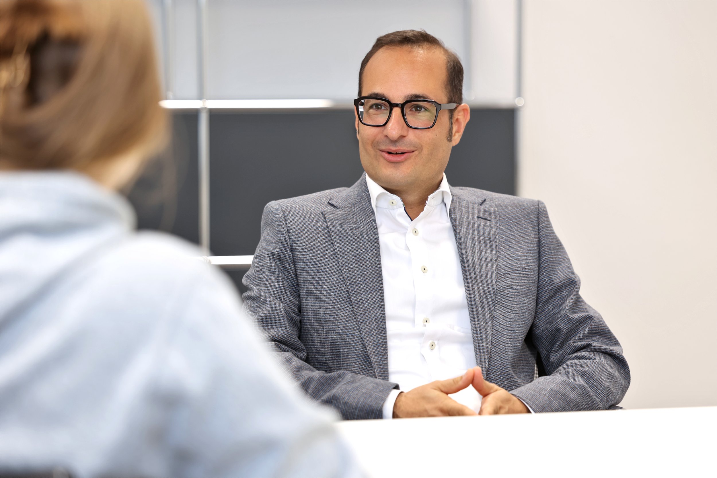 Herr Rokni-Yazdi im Anzug und mit Brille sitzt an einem Tisch und spricht mit einer Mandantin, die unscharf im Vordergrund ist. Es ist ein Beratungsgespräch im Büro zu sehen.