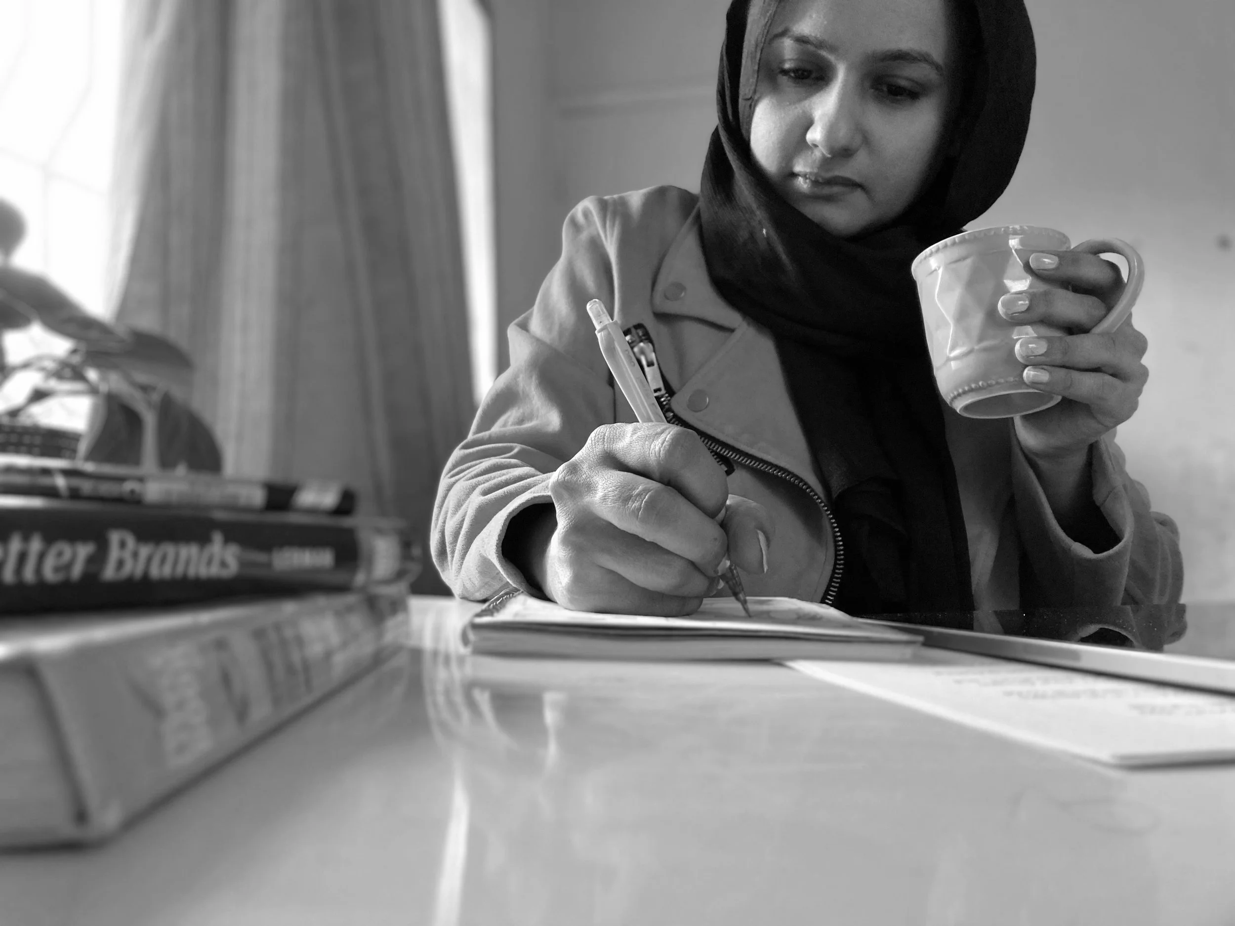 A woman wearing a hijab and a jacket, sitting at a table, writing in a notebook and holding a mug, with books and papers on the table.