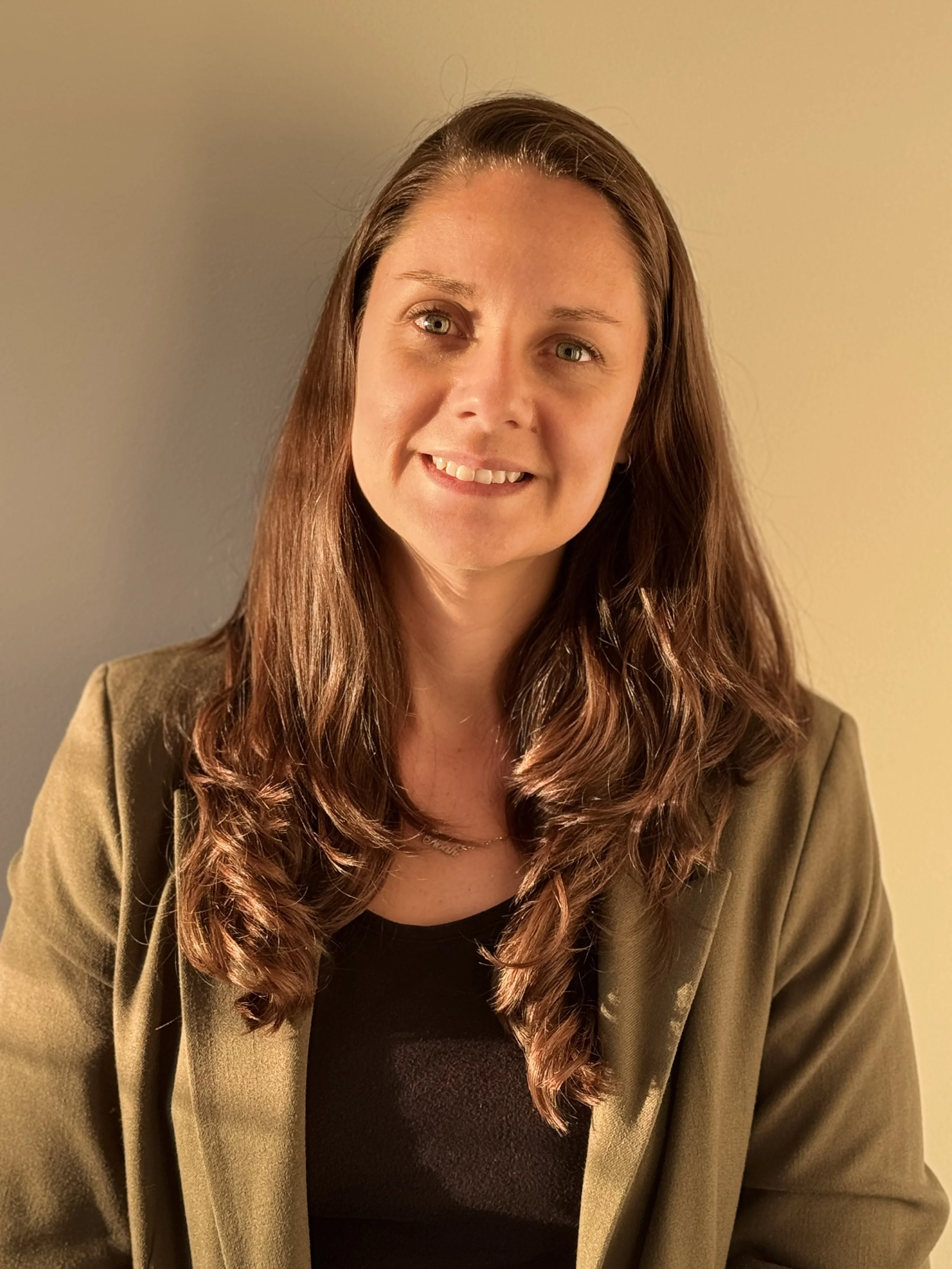 A woman with long brown hair, wearing a khaki blazer and black top, smiling softly at the camera against a plain, beige background.