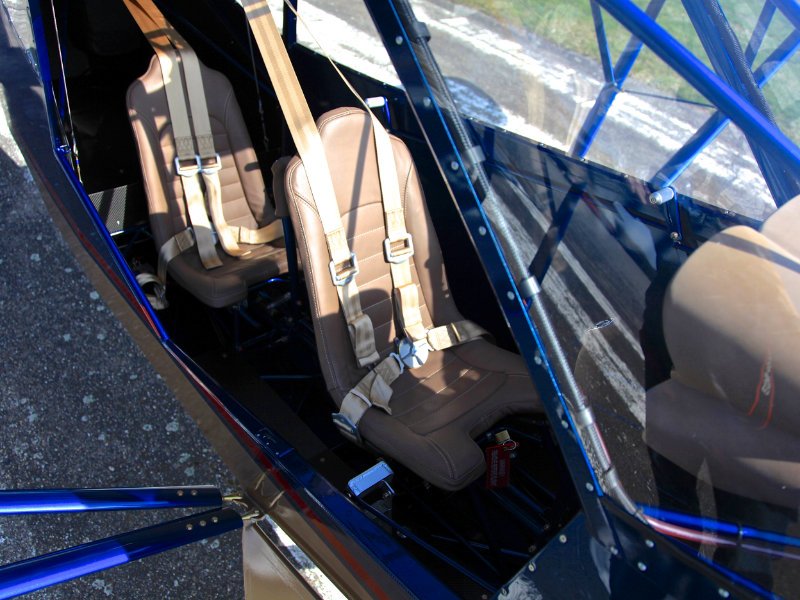 Interior of a lightweight racing car with two black racing seats, black harnesses, and a black carbon fiber interior. The rear panel displays the 'Blackwing Sweden' logo.