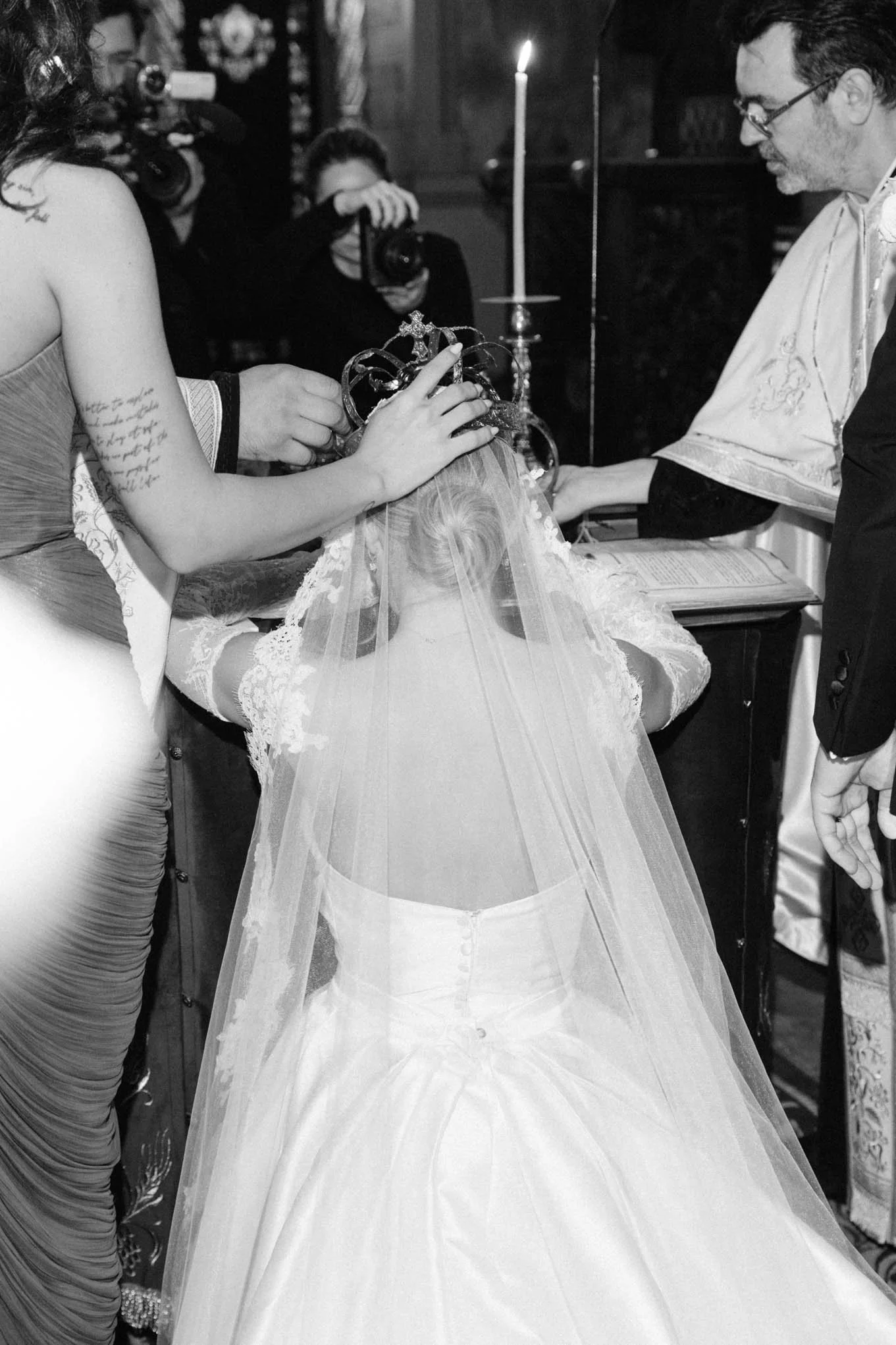 A bride kneeling during a wedding ceremony as a priest places a crown on her head, with women and photographers observing. French style classic wedding! 
