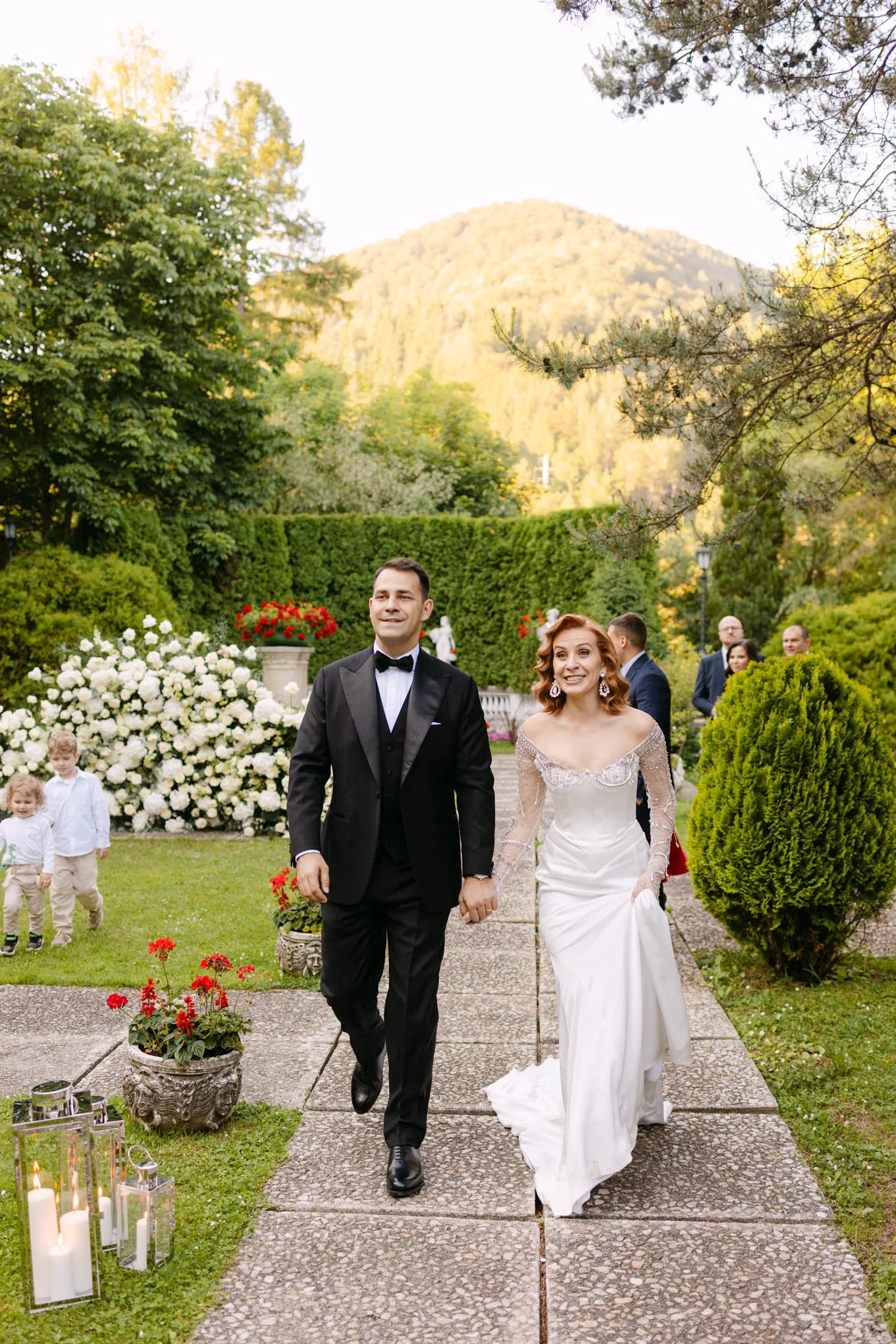 A couple dressed in wedding attire, holding hands and walking outdoors on a wedding day, with a lush green garden, flowers, and mountain scenery in the background. Like Tuscany style Villa garden.