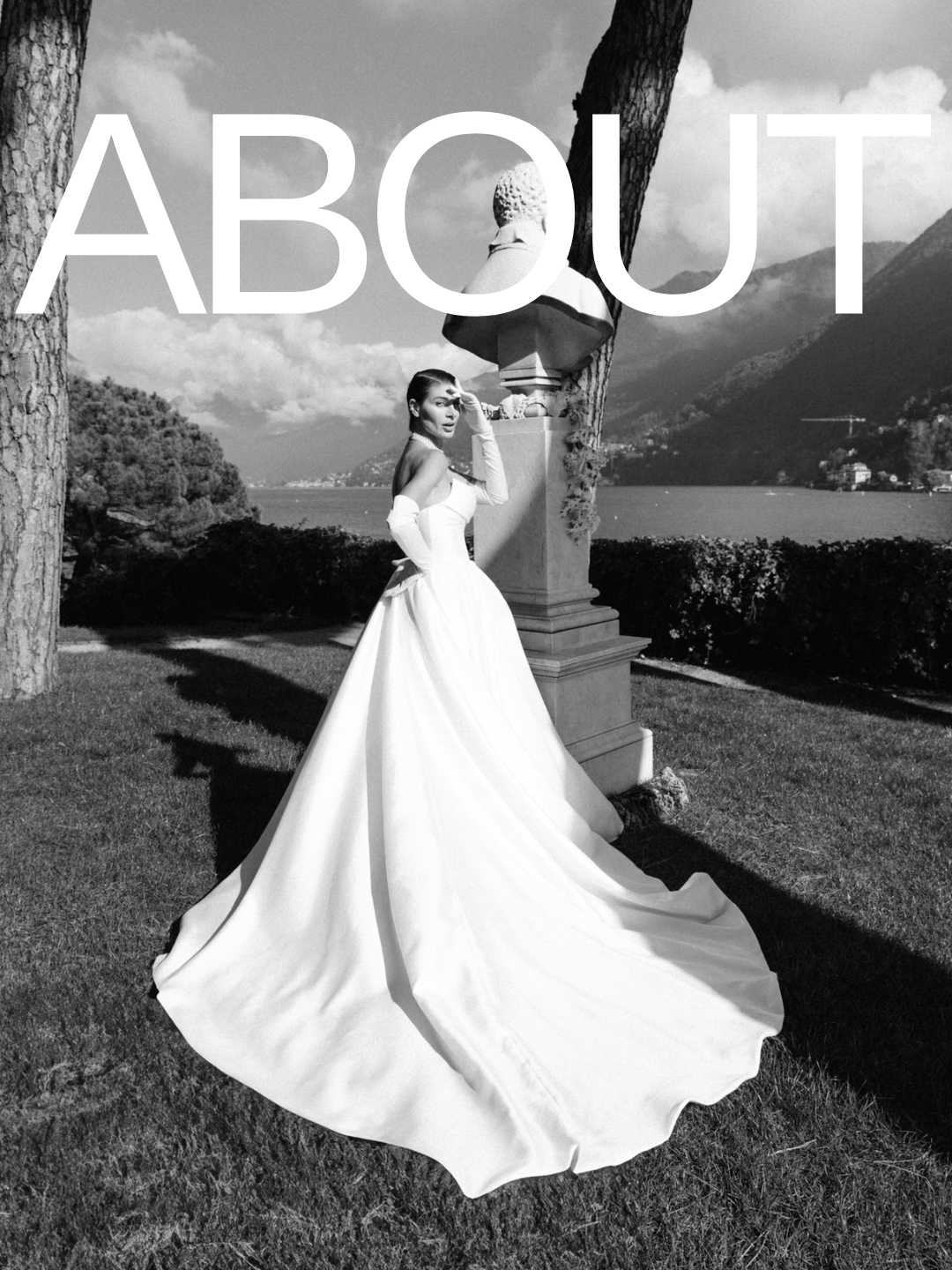 A woman in a wedding dress standing outdoors in front of a statue, with water and mountains in the background.