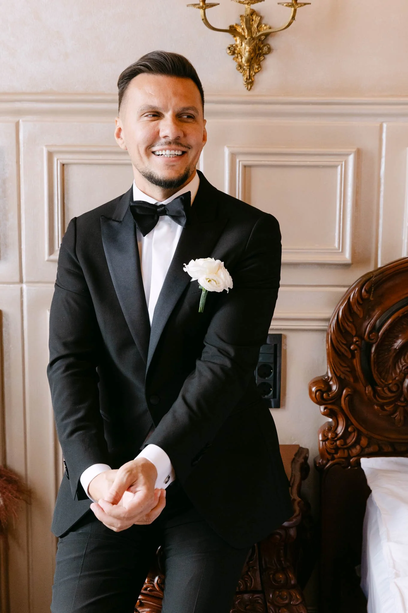 A man in a tuxedo with a white flower boutonniere, smiling and sitting on a wooden ornate chair in a room with cream paneled walls and a gold wall sconce. As seen in vogue!