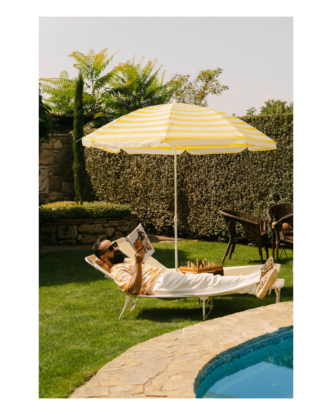 Man relaxing on a lounge chair by a pool, reading a magazine, shaded by a yellow and white striped umbrella, with lush green plants and chairs in the background.