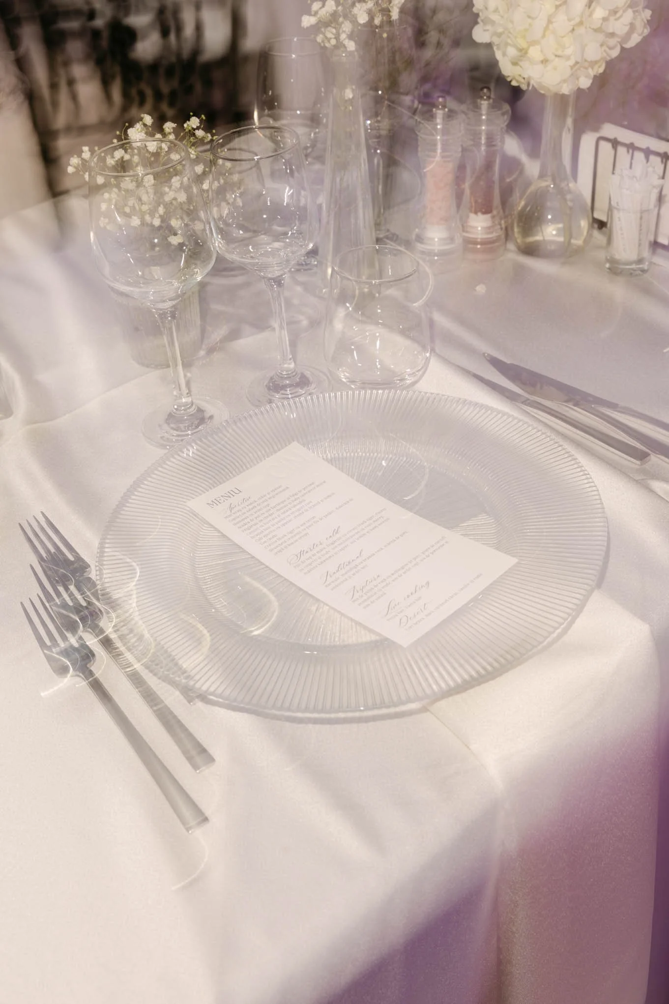 A formal table setting with a clear glass charger plate, three forks on the left, a knife on the right, a menu on the plate, three wine glasses, a water glass, and decorative flowers in vases in the background. Vogue style!