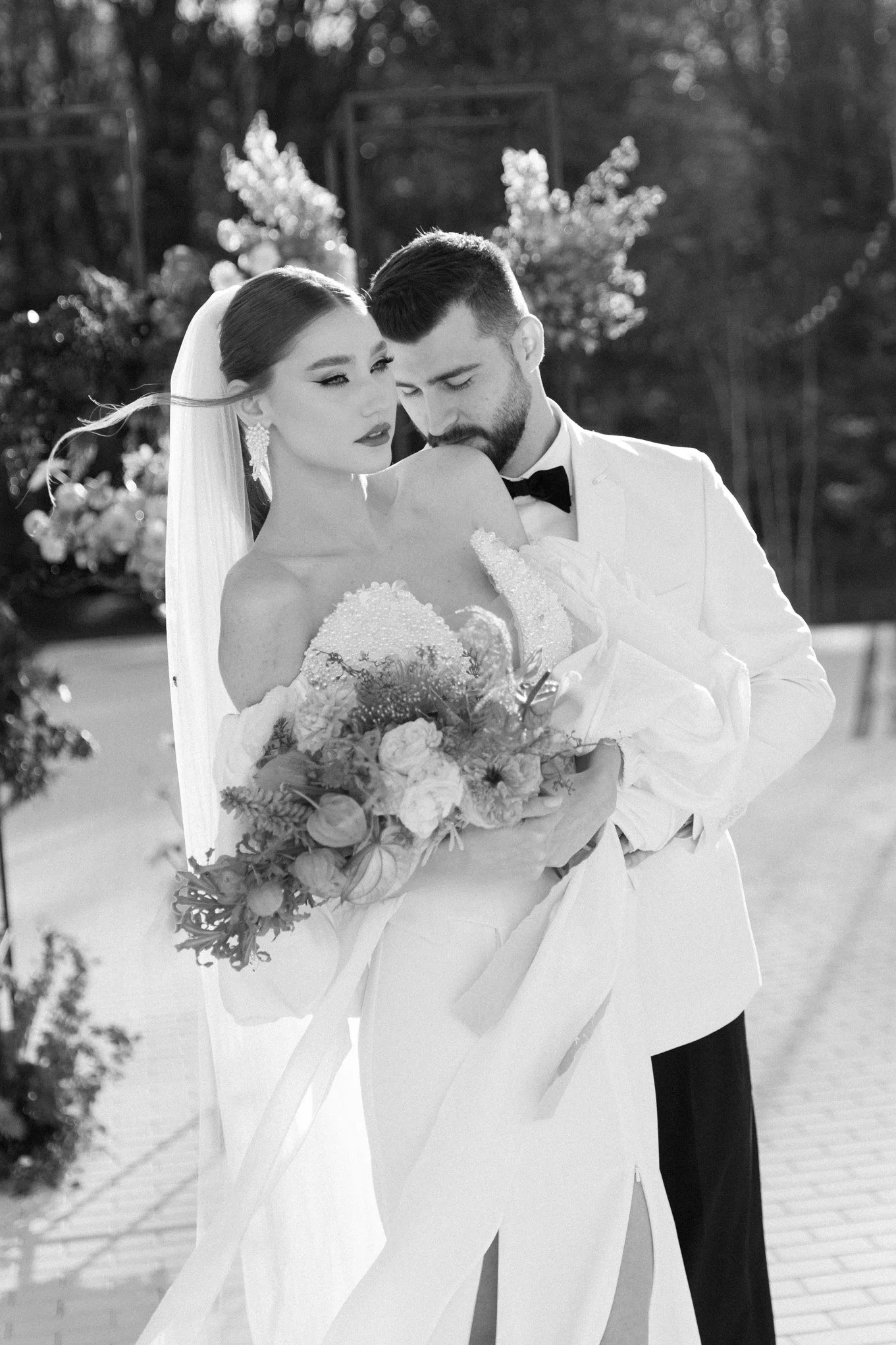 A bride and groom in wedding attire embrace outdoors, with the groom kissing the bride's shoulder. The bride holds a bouquet of flowers, and the scene is misty and romantic.