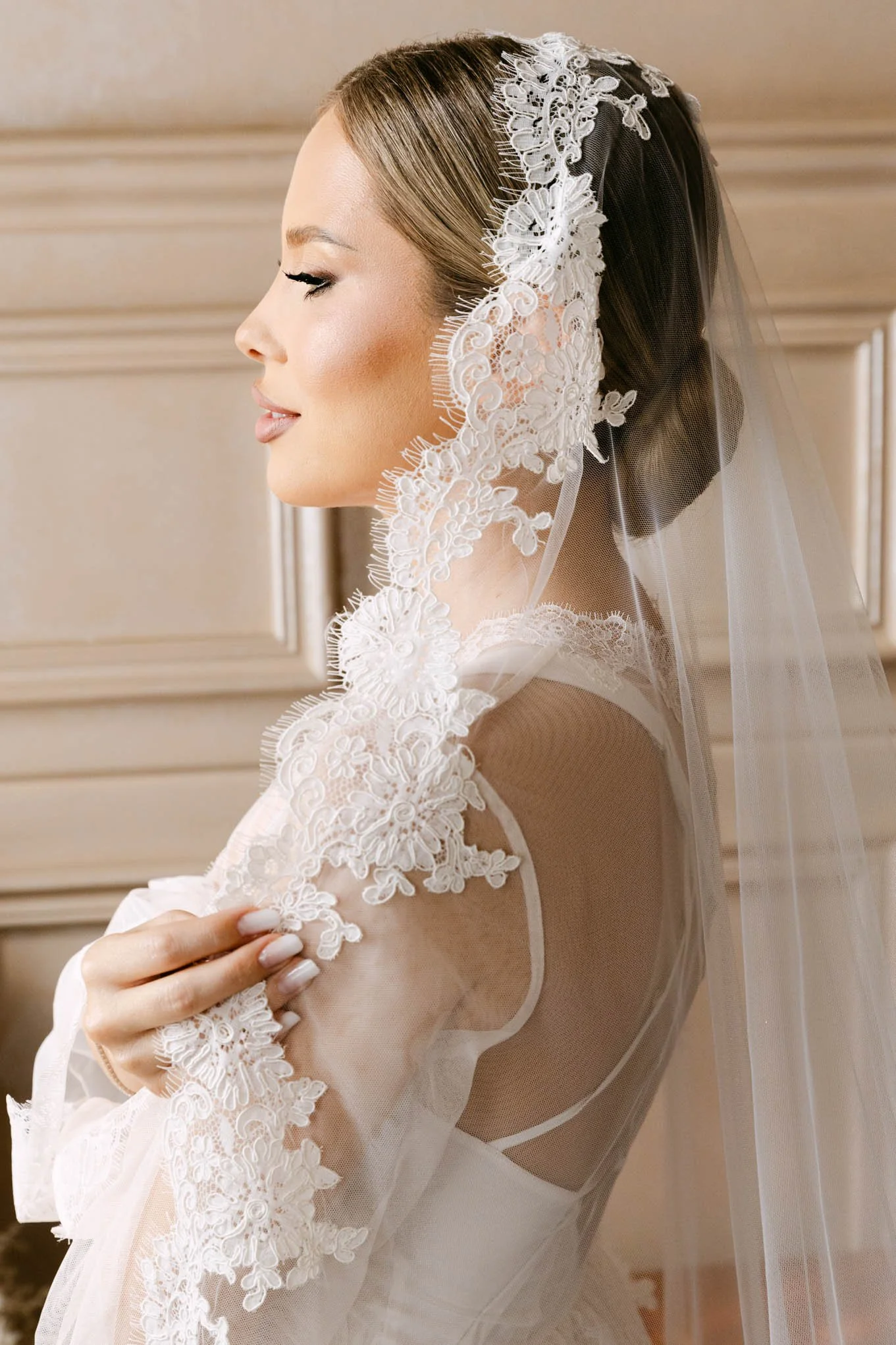 A bride with light brown hair in a bun, wearing a wedding dress and veil, holding her arm with lace floral details. She is standing indoors with beige walls. Vogue Style!