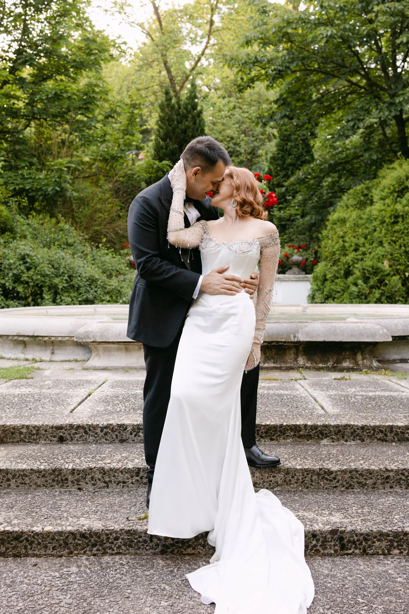 A bride and groom share a kiss in a lush, green outdoor  Tuscany garden  style with a fountain in the background.