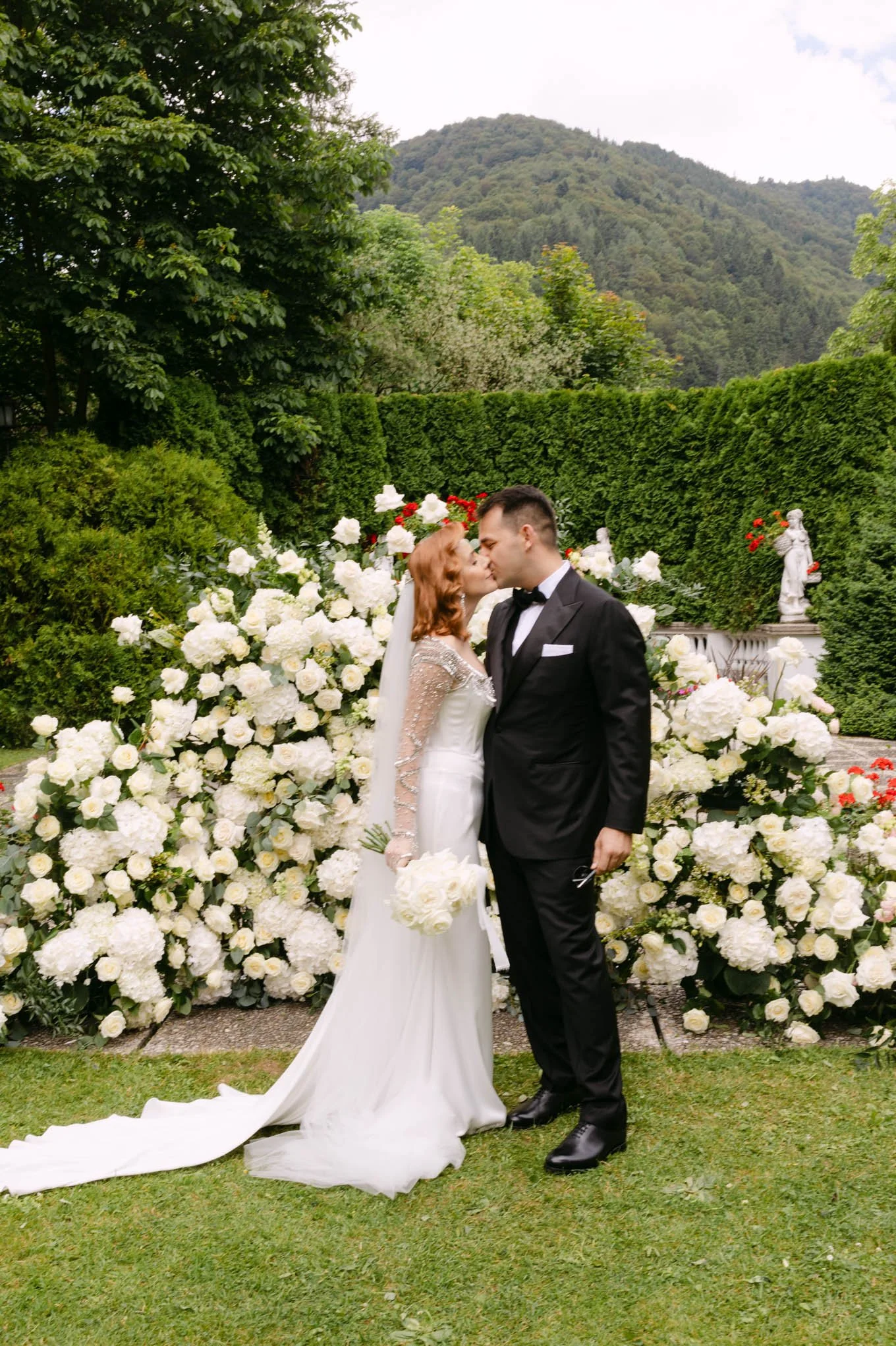 A bride and groom kiss in tuscany style  garden with lush greenery and mountains in the background, surrounded by white floral arrangements.